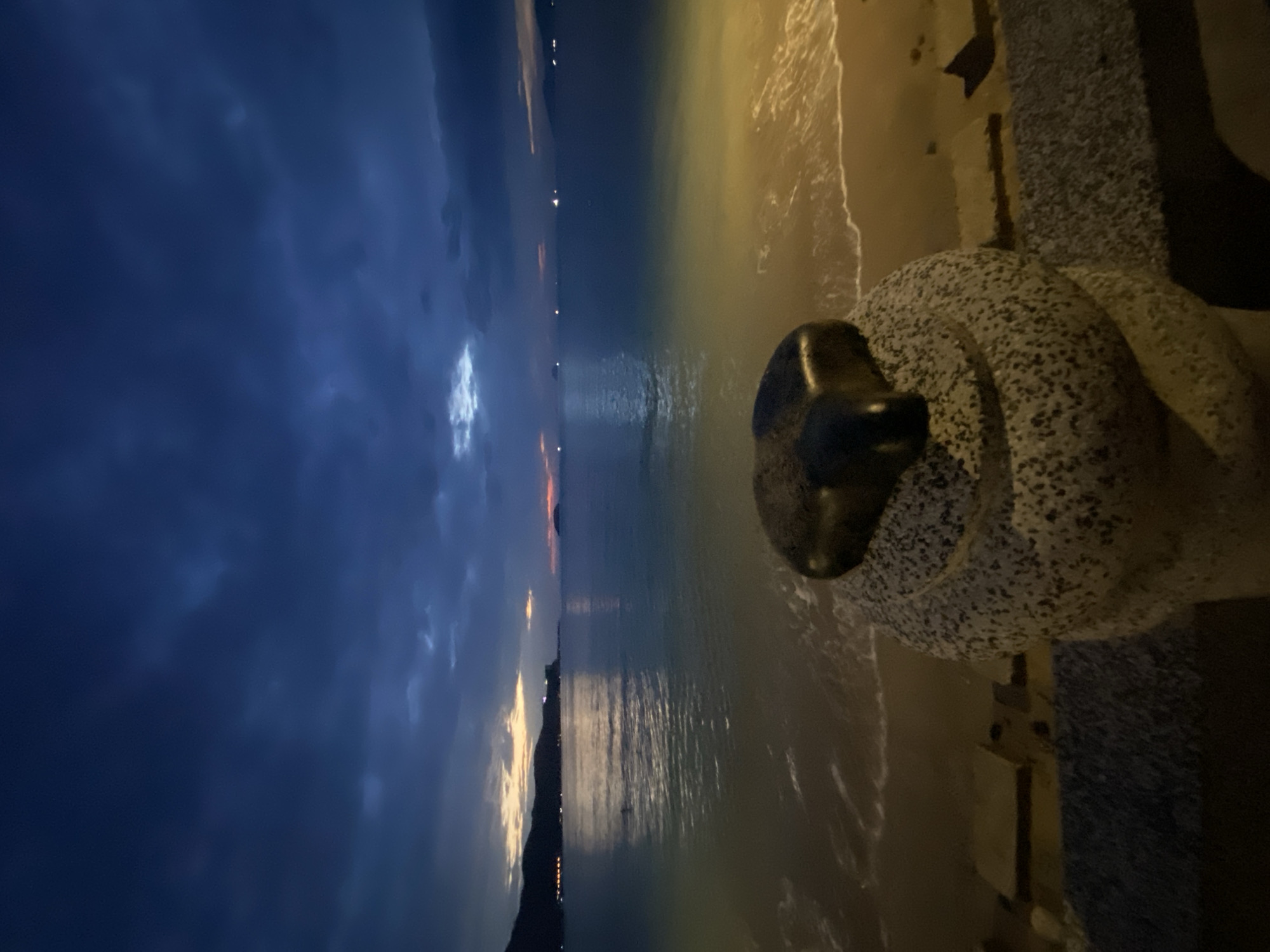 A photograph captures a twilight scene at a beach in Nha Trang, Vietnam. In the foreground, a textured stone pedestal topped with a dark, heart-shaped object is situated near the water's edge. The ocean stretches towards the horizon, reflecting the fading light of the sky. Distant hills and lights from the city are visible along the coastline. The sky is a deep blue with dramatic clouds, hinting at the transition from day to night. Gentle waves lap the sandy shore, creating a serene and atmospheric view.