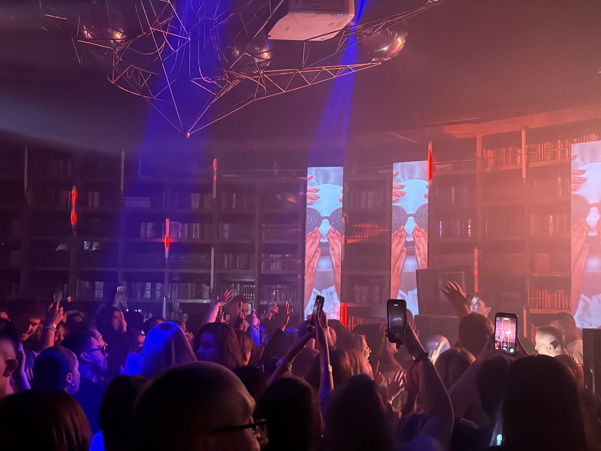 The image depicts a crowded event in what appears to be a nightclub or concert venue. Many people are visible, their faces and bodies mostly obscured by the dim lighting and density of the crowd. Some individuals are holding up smartphones, presumably recording or photographing the performance or event. The background features large video screens displaying abstract images, and decorative elements that resemble bookshelves, suggesting a themed or unique interior design. Above the crowd, complex lighting fixtures and disco balls are suspended. The lighting is primarily blue and red, creating a vibrant and energetic atmosphere. Based on the style of the event and available information, the location is likely a club or venue in Moscow, Russia.