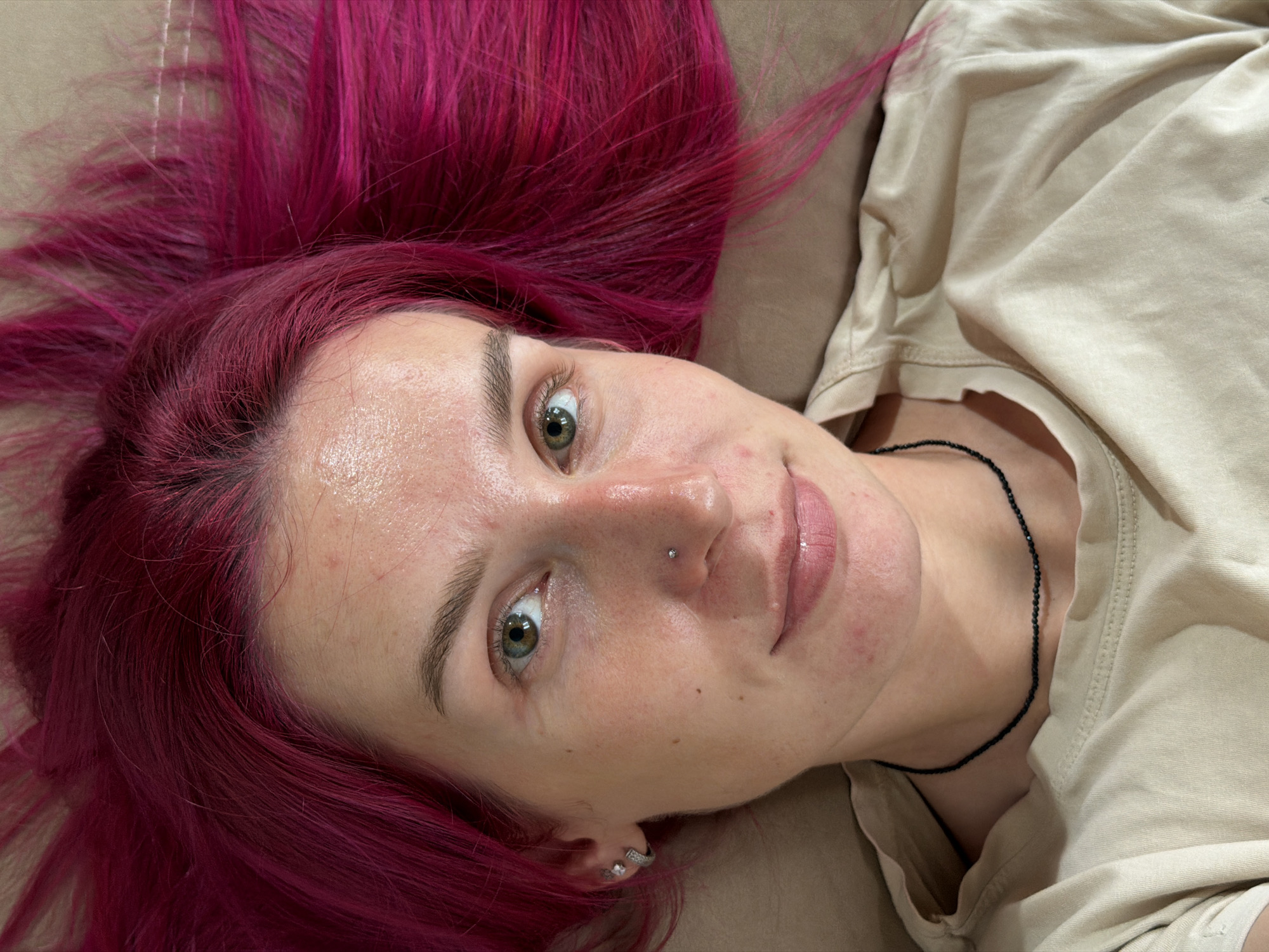 The image shows a close-up, overhead shot of a young woman with vibrant pink hair lying down. She is looking directly at the camera with a relaxed expression. Her eyes are open and a light green color. She has a subtle smile on her lips, which appear to be slightly glossy. She is wearing a light-colored t-shirt and a black beaded necklace. A small silver stud is visible in her nose, and she has a hoop earring in her left ear. Her hair is spread out around her head, creating a vibrant frame. The background appears to be a textured surface, possibly a bed or sofa. The overall lighting suggests an indoor setting. There is no discernible indication of the city of Chaikovsky, Russia, within the image content itself.