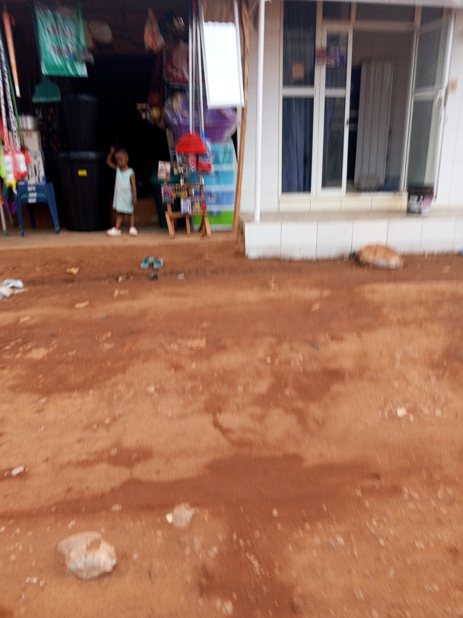 A dirt road in Abuja, Nigeria, is in the foreground. A store with a child standing inside is on the left side of the image. To the right of the store is a building with white framed windows.