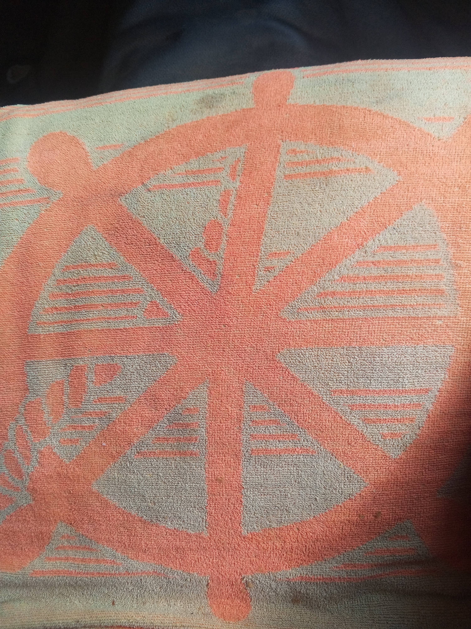 The image shows a close-up, overhead view of a towel with a circular, sunburst-like pattern. The pattern is rendered in a coral pink color against a pale blue or gray background. The design consists of a central circle from which eight rays emanate outwards. Each ray is divided into three sections, with the outermost sections filled with horizontal lines. The towel appears to be made of terry cloth, with a textured surface. There are some dark specks on the towel, possibly dirt or stains, particularly noticeable on the right side of the image. The lighting suggests it might be indoors or in a shaded outdoor area, as there is a dark shadow obscuring the right edge of the towel. No text is visible. The overall impression is of a decorative towel, possibly used for beach or bath. The location context provided is Altagracia, Venezuela, which could suggest it is a towel used in that region, but there are no specific visual cues within the image to confirm this. The scene is static, focusing solely on the towel's pattern and texture.