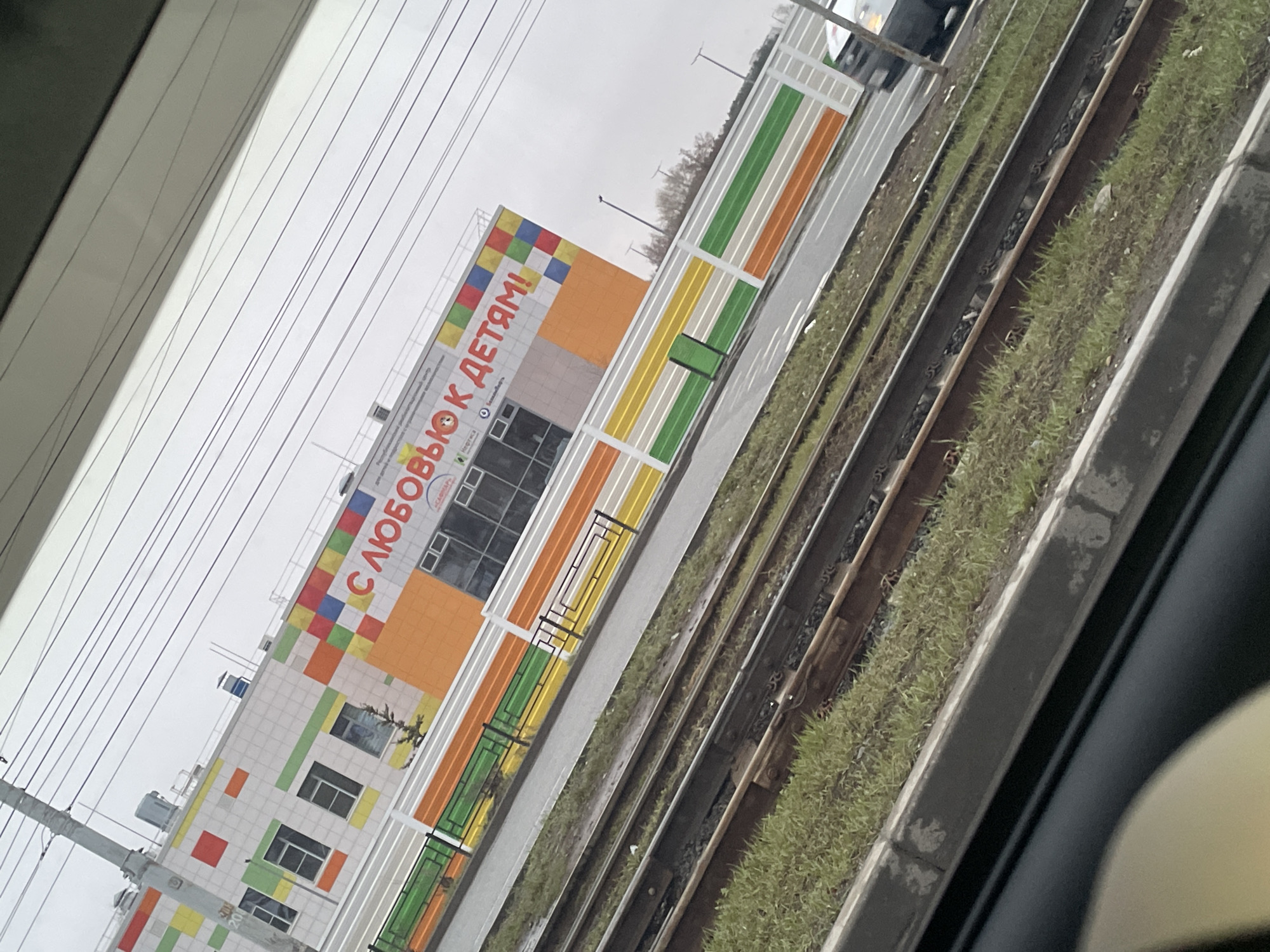 The image shows a building with a sign in Russian that reads "С любовью к детям!" (With love to children!). The building has colorful square patterns on its facade. Adjacent to the building are train tracks with grass growing alongside them. The photograph appears to be taken from inside a vehicle, looking out towards the building and train tracks. Based on the building's architectural style and the Russian text, the location is likely in Russia. Additional context: The architecture of the building indicates that it's a kindergarten. The location can be narrowed down to Izhevsk, Russia because the kindergarten in the image is located there.