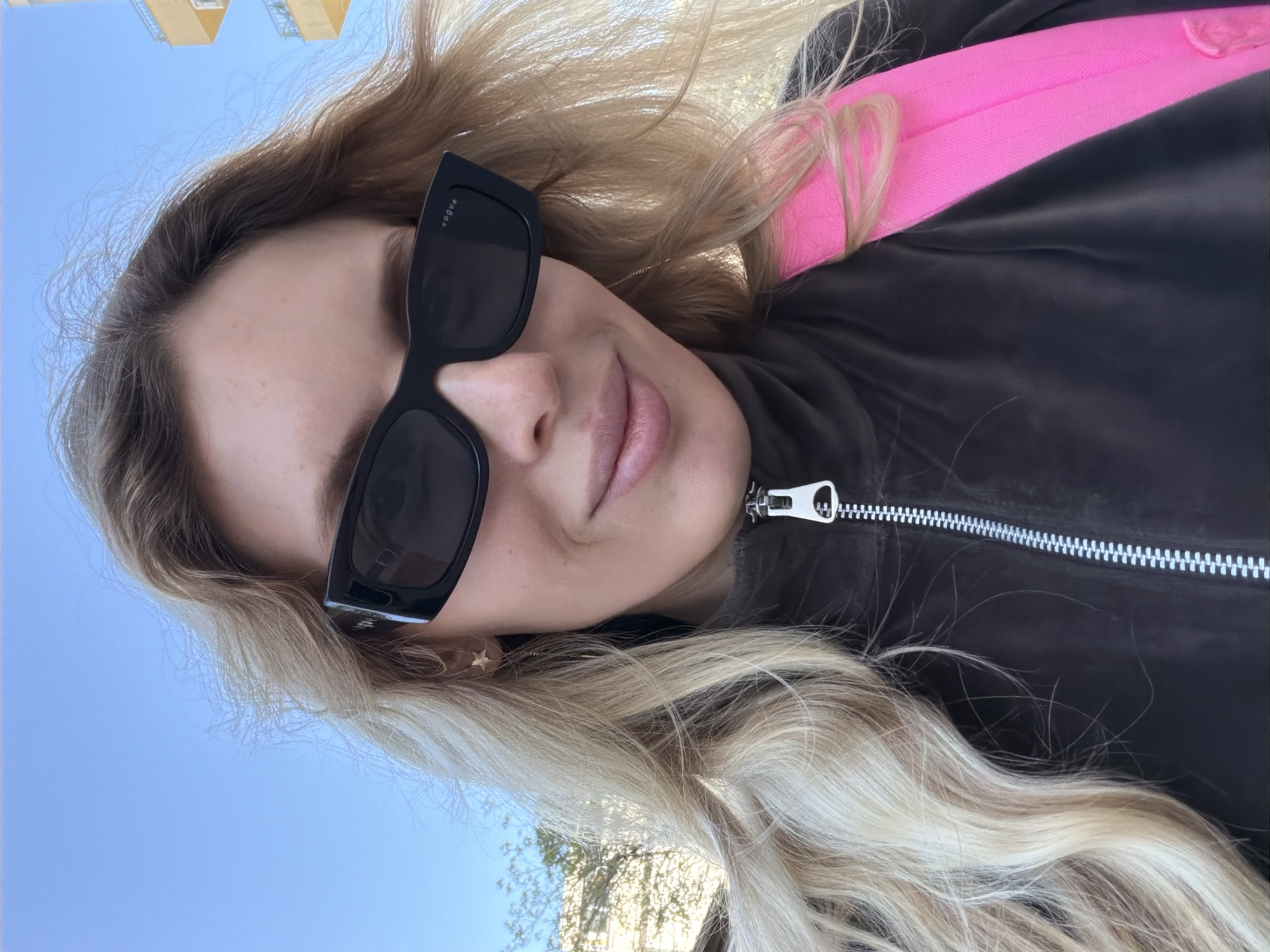 The image is a portrait-style shot of a woman, rotated 90 degrees clockwise, giving the impression of her lying down or the photo being taken at an unconventional angle. The woman, who appears to be in Budva, Montenegro, is seen from the chest up, with long, wavy blonde hair cascading around her. She wears large, rectangular black sunglasses, with the word "VOGUE" visible in white text on the temple of the left lens. Her lips are slightly parted, revealing a subtle, relaxed expression.
She is dressed in a dark, possibly velour or velvet, zippered jacket. A prominent silver zipper runs down the front of the jacket. Over her left shoulder, a vibrant pink strap or portion of a bag is visible, adding a pop of color to her attire. A small, delicate gold star earring is visible in her right earlobe.
The background consists primarily of a clear, bright blue sky, indicating sunny weather. In the upper left corner, parts of a light-colored building, possibly yellow, are visible against the sky. Towards the lower right, faint shadows or foliage from trees can be seen, suggesting an outdoor setting. The bright lighting points to the photograph being taken during the daytime. The overall impression is a casual, outdoor selfie.