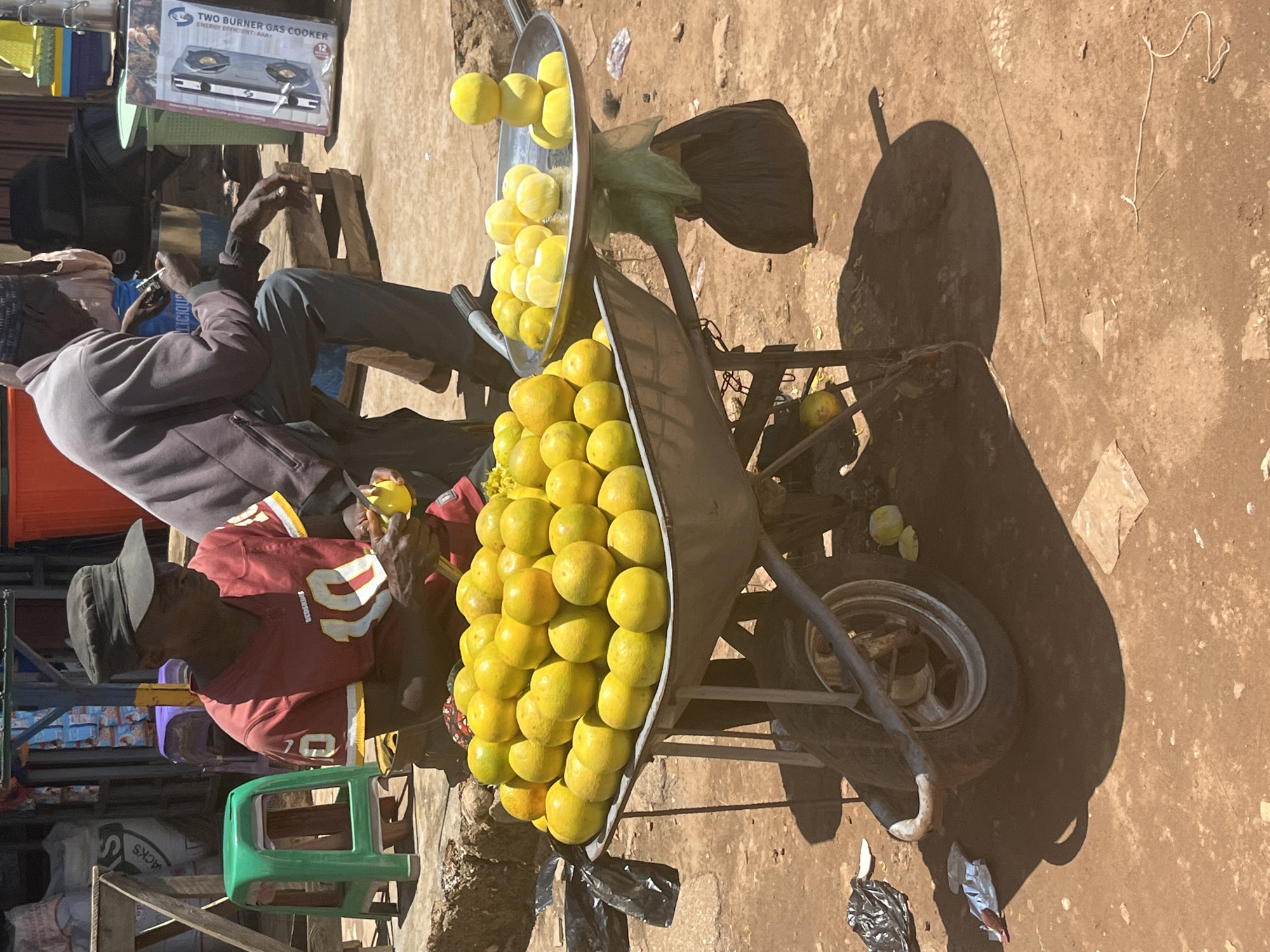 The image shows a street vendor in the city of Jos, Nigeria, selling oranges. A man wearing a dark cap and a red and white football jersey is peeling an orange. He is sitting next to a wheelbarrow filled with oranges. Another man, wearing a gray jacket and dark pants, is seated in the background, also peeling an orange. The scene is set on a dusty, outdoor market stall with other vendors and goods visible in the background, including a gas cooker box. The overall lighting suggests it is daytime, likely sunny.