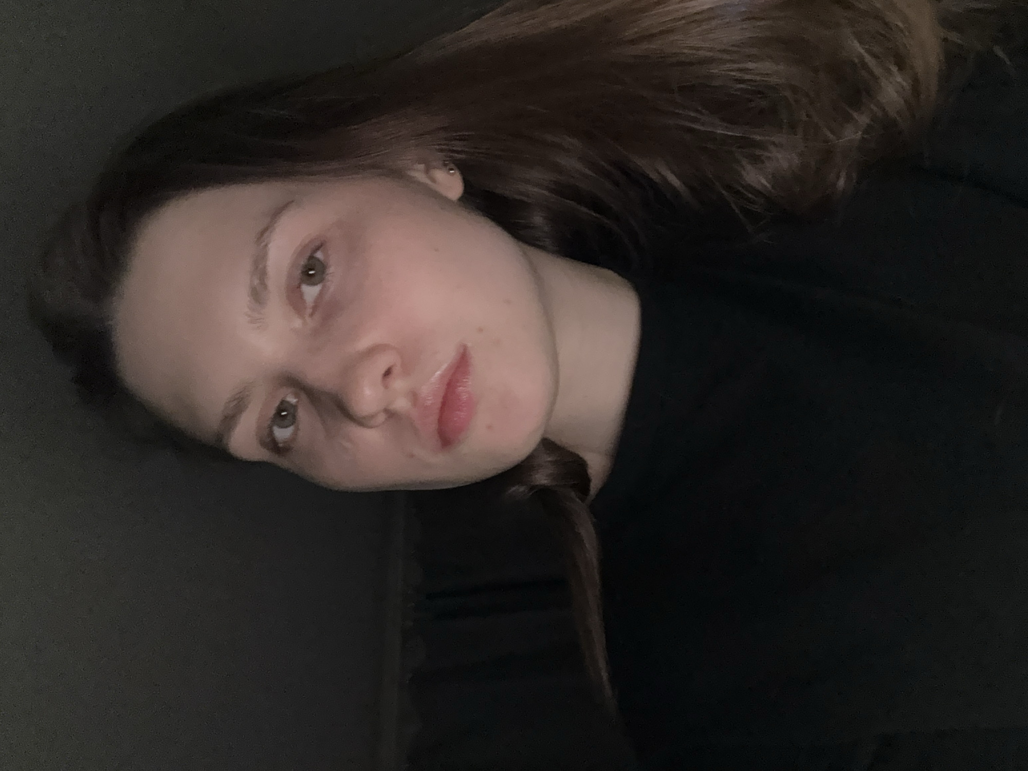 A person is angled with their head towards the upper left corner of the frame. They have light skin, brown hair, and their lips are slightly glossy. The person is wearing a dark-colored top. The background is a dark gray. The location is not discernible from the image. The location is not Saint Petersburg, Russia.