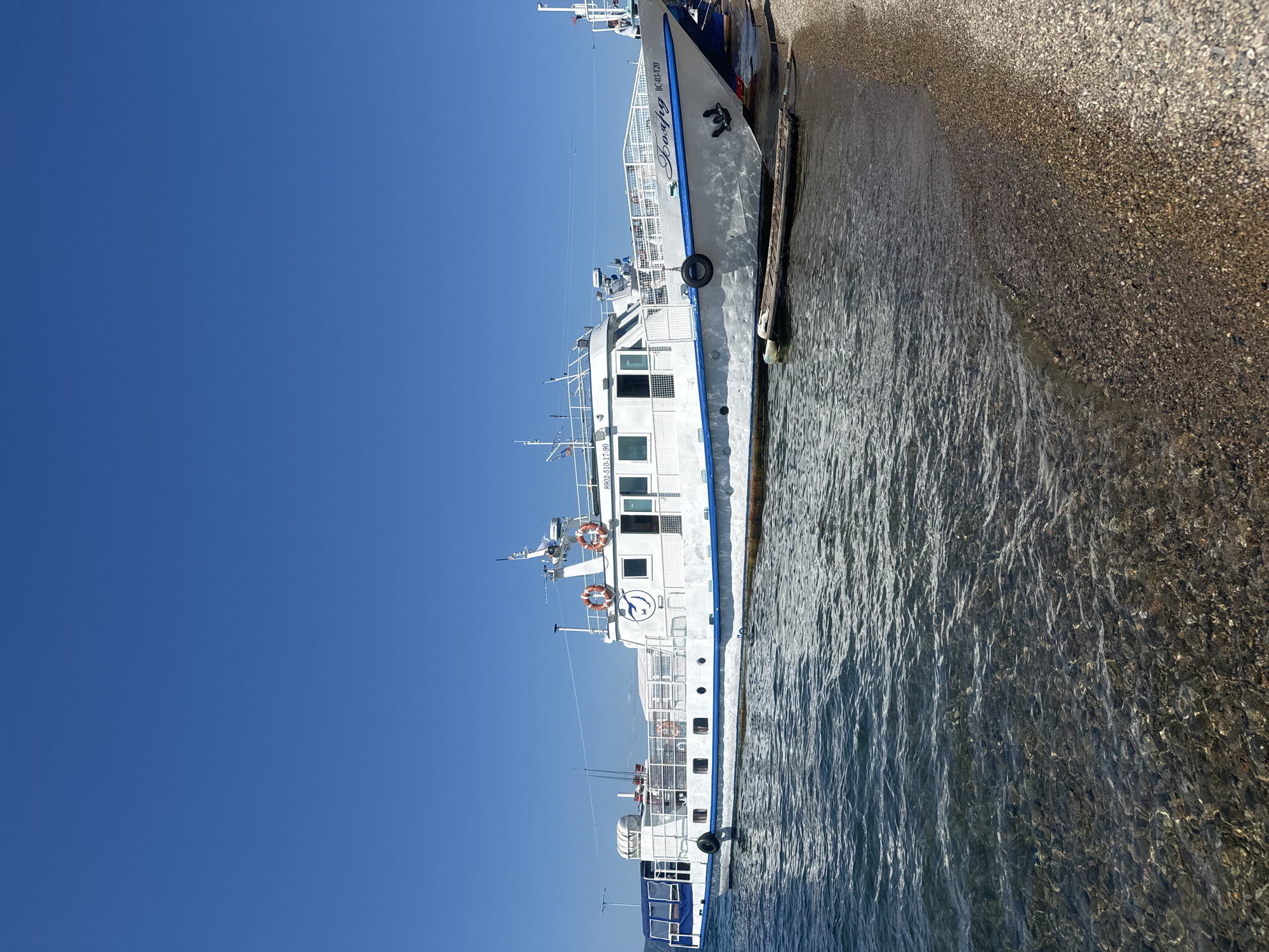 A white passenger boat with blue trim is docked on a pebble beach under a clear blue sky. The boat has several windows, life preservers, and a name written on its side in Cyrillic script. The water is calm and clear, with the seabed visible. In the distance, land can be seen. The scene appears to be a sunny day in Listvyanka, Russia, likely at the shore of Lake Baikal, given the characteristic pebble beaches and the type of boat.