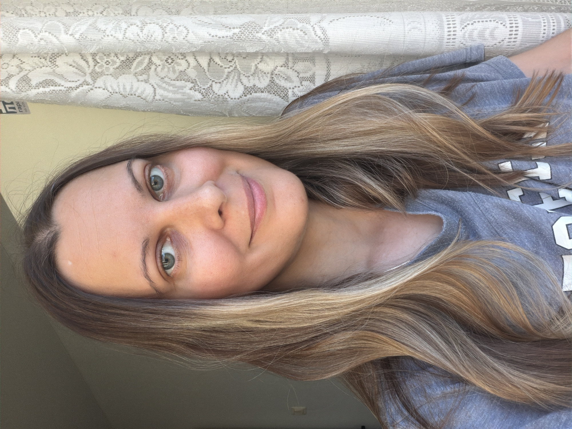 A woman with long, wavy, blonde and brown hair is posing for a selfie in what appears to be a home environment. She is wearing a grey t-shirt. Behind her, to the right, is a white lace curtain. The background to the left is a plain wall. There is a small white mark on her forehead. She is looking directly at the camera with a slight smile. The image is a close-up, focusing on her face and upper torso. There is no information in the image to suggest the location is Izhevsk, Russia.