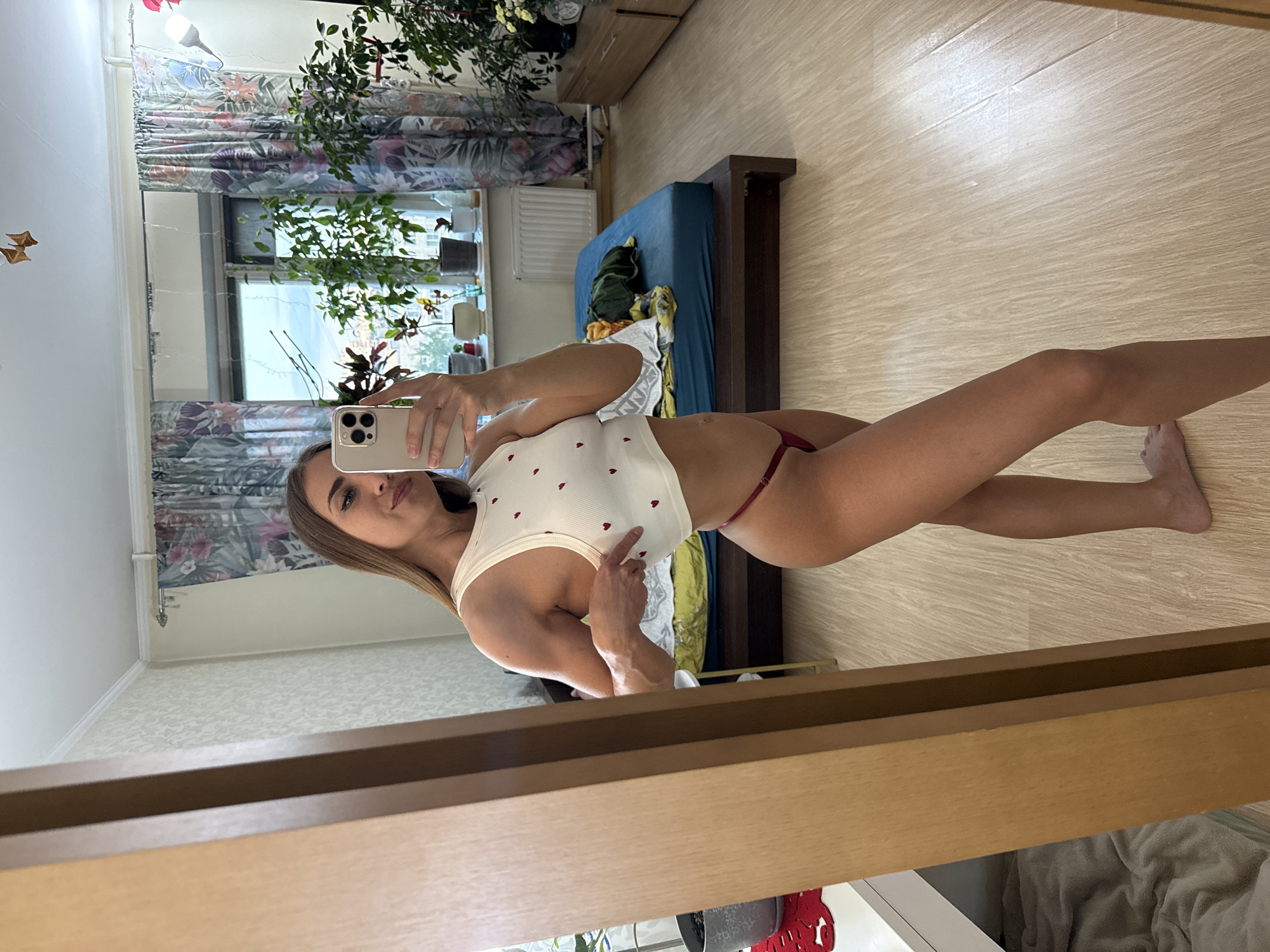 A woman is taking a selfie in a mirror in what appears to be a bedroom. She is wearing a white tank top with small red hearts and red underwear. The room has light wood laminate flooring and a light-colored wall. In the background, visible in the mirror, is a window with floral curtains, some plants, and a wooden bed frame with a blue mattress. There is also a dresser or cabinet with items on it. The lighting suggests it is daytime. The woman is looking at the camera and has a neutral expression.