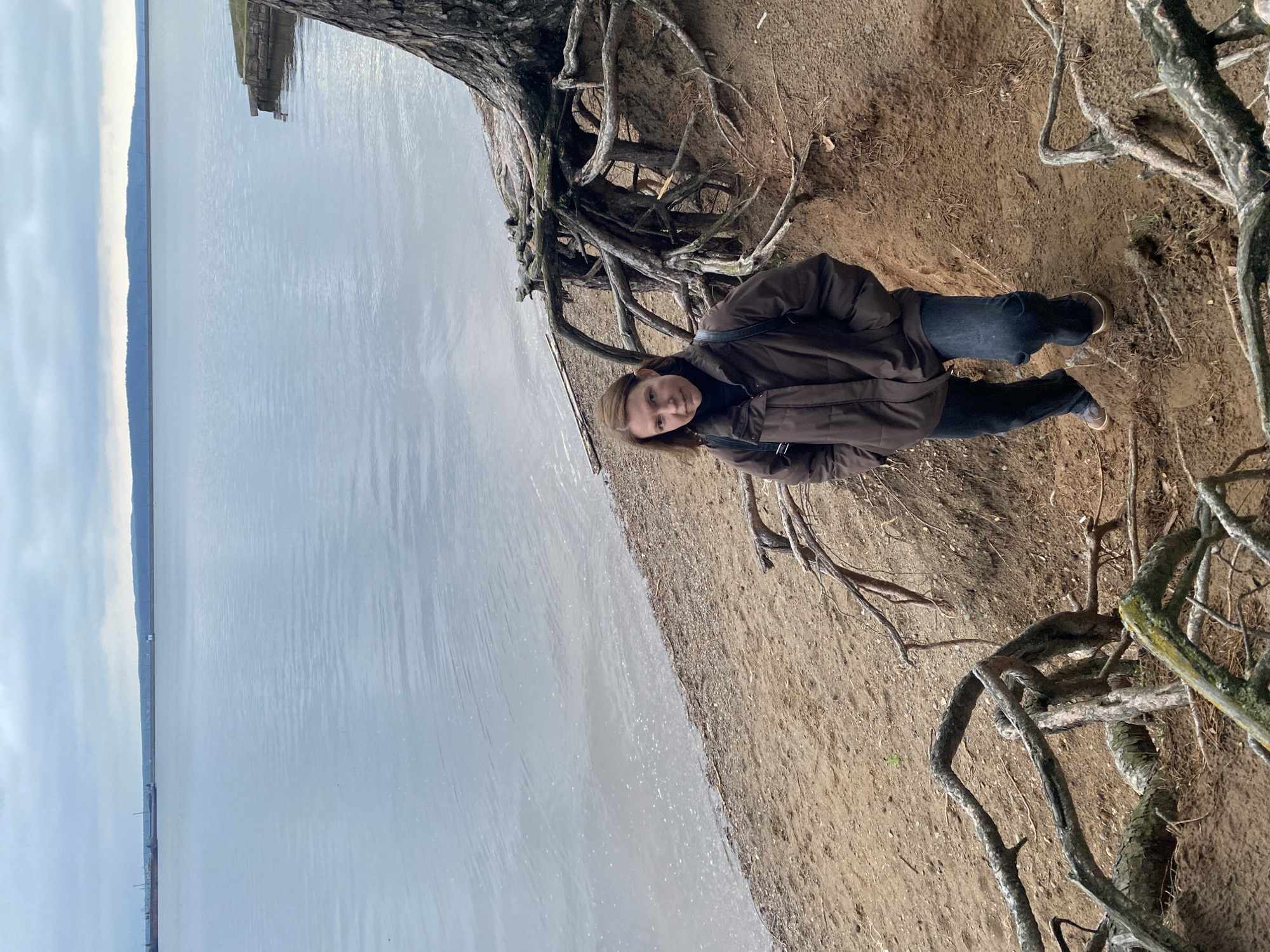 A woman stands on a sandy bank next to a body of water under a large tree with exposed roots. She is wearing a dark brown jacket, dark blue jeans, and tan shoes. The water is gray and still, with land visible in the distance under a cloudy sky. The sandy bank is brown and covered with tree roots and branches. The location is Chaikovsky, Russia.