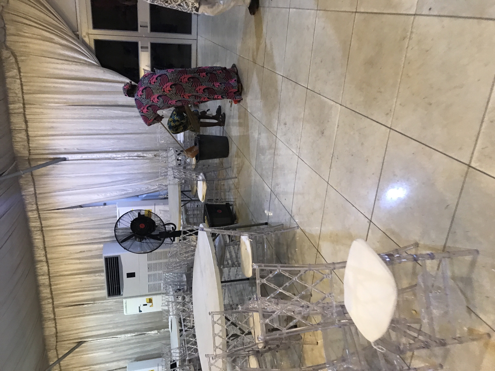 The photograph captures an interior scene in Oko Erin, Nigeria. The setting appears to be within a large tent-like structure, evidenced by the draped fabric forming the walls. The floor is tiled with light-colored, square tiles.
A person wearing a colorful, patterned outfit is mopping the floor with a mop and bucket. Tables with white tablecloths and transparent chairs with white padded seats are set up, seemingly arranged for an event. An industrial fan and an air conditioning unit are visible, mounted on the tent structure. The lighting is bright, with some areas casting shadows. The general impression is that of event preparation or post-event cleanup.