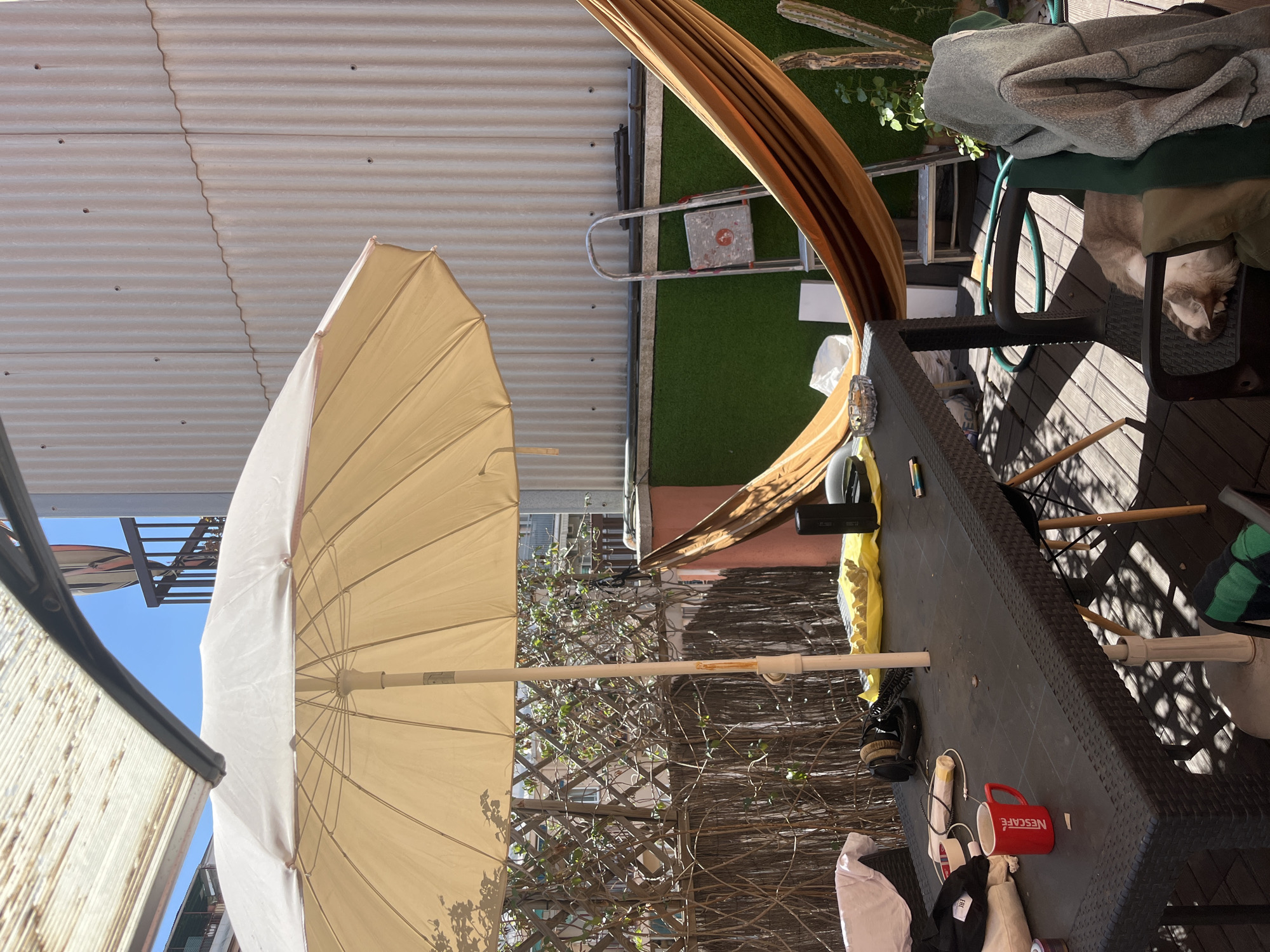 The image shows an outdoor patio setting in Barcelona, Spain. A large beige umbrella is prominent, partially obscuring a lattice fence with foliage behind it. A brown hammock is strung between two supports, casting shadows on the scene. A dark-colored patio table is cluttered with various items, including a red "NESCAFÉ" mug, clothing, shoes, and electronic accessories. A ladder and artificial turf are visible in the background. A cat is resting in a chair. The angle suggests the photo was taken from a low perspective. Corrugated metal siding is visible at the top of the frame.