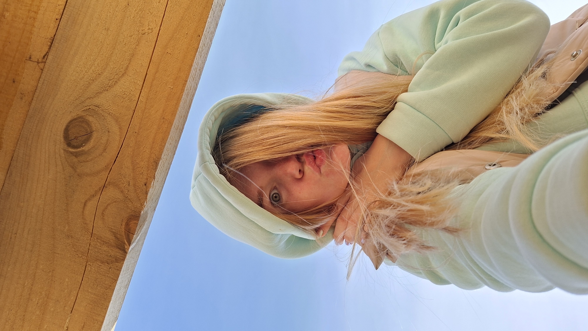 A low-angle, upward-facing shot captures a young woman in a light green hoodie, positioned in front of a clear blue sky and a section of wooden structure. Her blonde hair partially covers her face as she makes a puckered expression with her lips. The wooden beams show natural grain patterns. The location is Большой Букор, Russia.