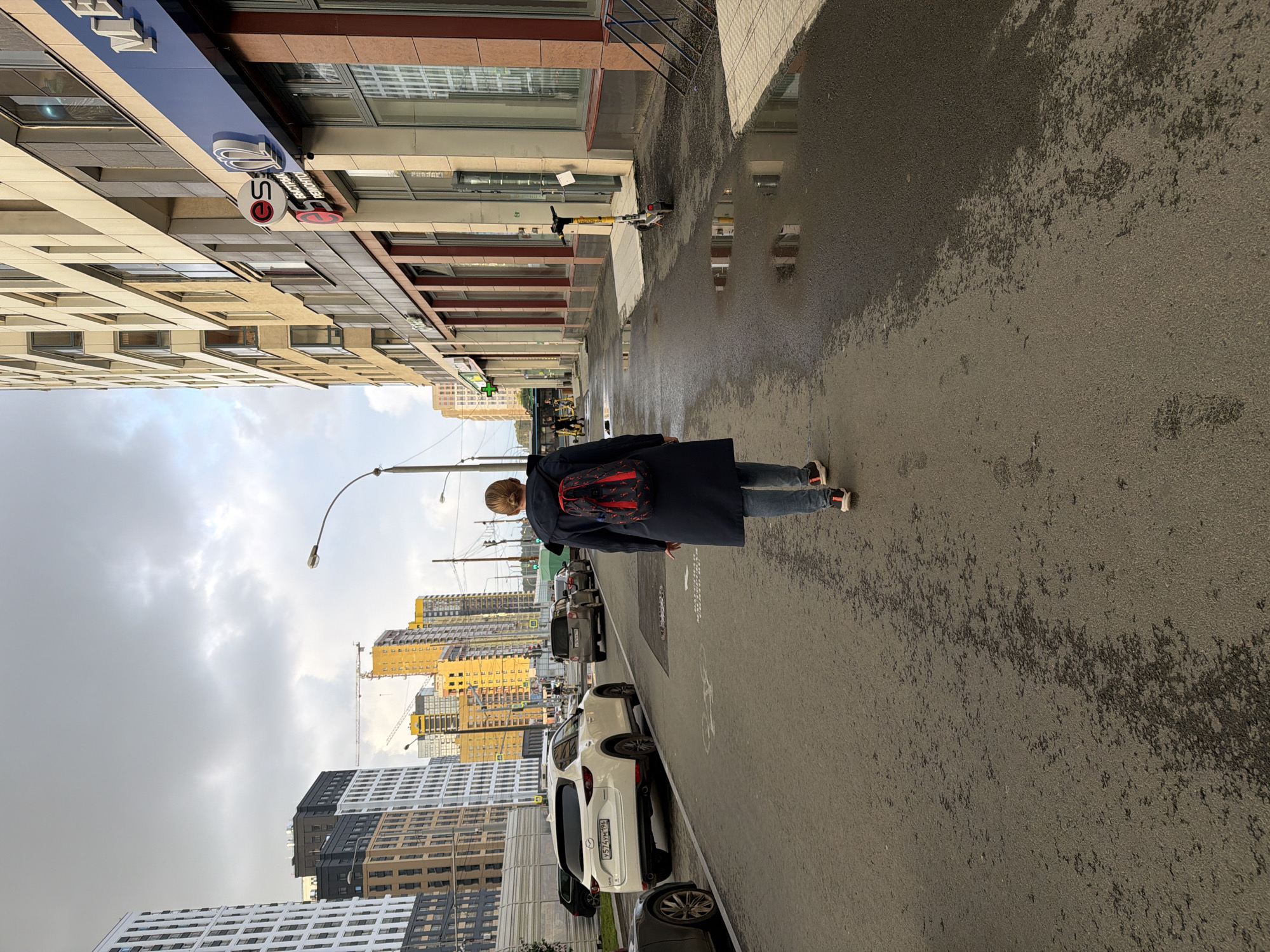 The image shows a person walking on a wet street in front of a modern building. The person is wearing a dark coat and blue jeans and has a backpack with red accents. The street has puddles reflecting the surrounding buildings. In the background, there are several multi-story buildings, some of which appear to be under construction, with cranes visible. The sky is overcast with clouds. The scene appears to be in an urban environment. Visible text includes "ES" on a circular sign on the building. The time of day is difficult to determine precisely due to the lighting conditions, but it appears to be daytime, possibly late afternoon or early evening given the overcast sky. The weather is wet, suggesting recent rain.