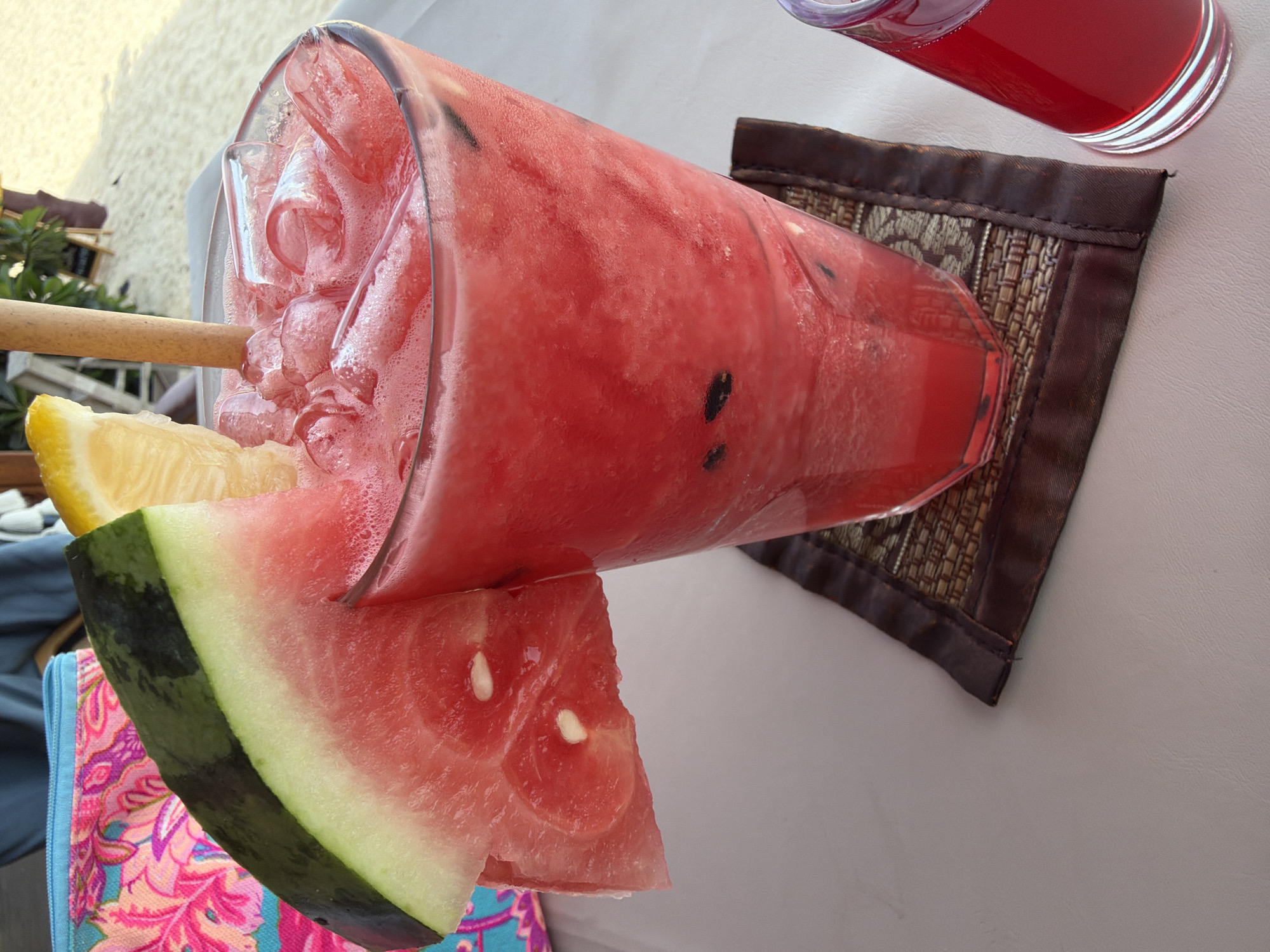 A close-up shot shows a tall, clear glass filled with a vibrant pink, icy beverage. A slice of watermelon with its green rind and red flesh is perched on the rim of the glass, with a wedge of lemon also adorning the top. A natural-colored straw extends into the drink, which is topped with frothy foam and ice cubes. The glass sits on a woven brown coaster. To the right, a smaller glass contains a similar red-hued liquid. In the background, a colorful fabric bag with a floral pattern is visible, and beyond that, a sandy beach and a hint of greenery suggest an outdoor setting, possibly in the city of Ko Samui District, Thailand, given the tropical ambiance.