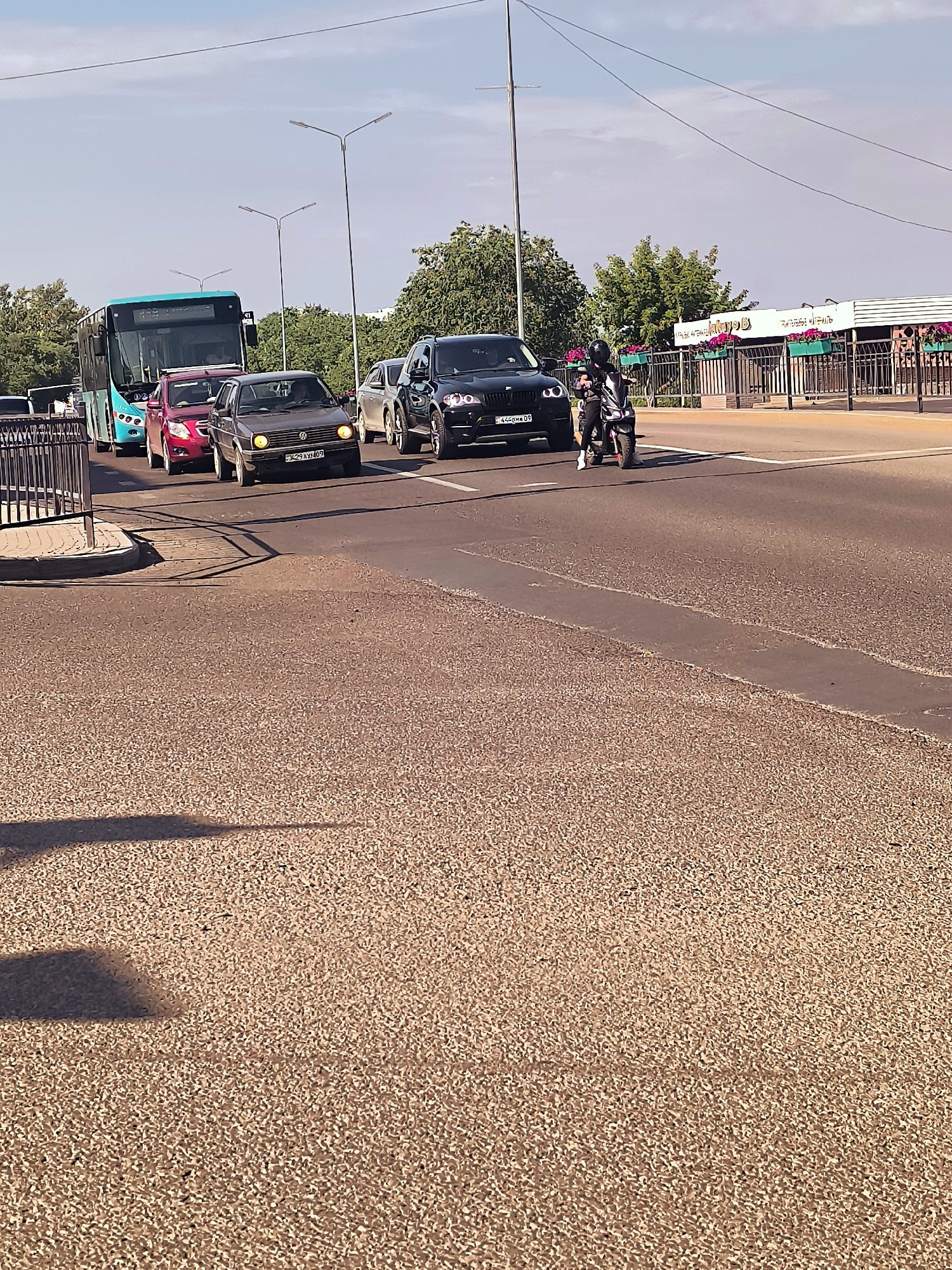 In Qarağandı city administration, Kazakhstan, a traffic scene unfolds on a sunny day. A turquoise bus is stopped in traffic, followed by a red car and a dark grey Volkswagen Golf. The Golf has its headlights on, and its license plate reads "K294AX09". Next to the Golf, a black SUV is also stopped, with its license plate showing "4260MM 09". A person on a motorcycle, wearing a helmet and dark clothing, is stopped to the right of the SUV, facing forward. In the background, there is a building with "Jalyn" written on it, possibly a restaurant or establishment, adorned with potted pink flowers along a railing. Trees and streetlights are also visible, contributing to the urban environment. The road surface is asphalt with a textured, gravelly appearance in the foreground.