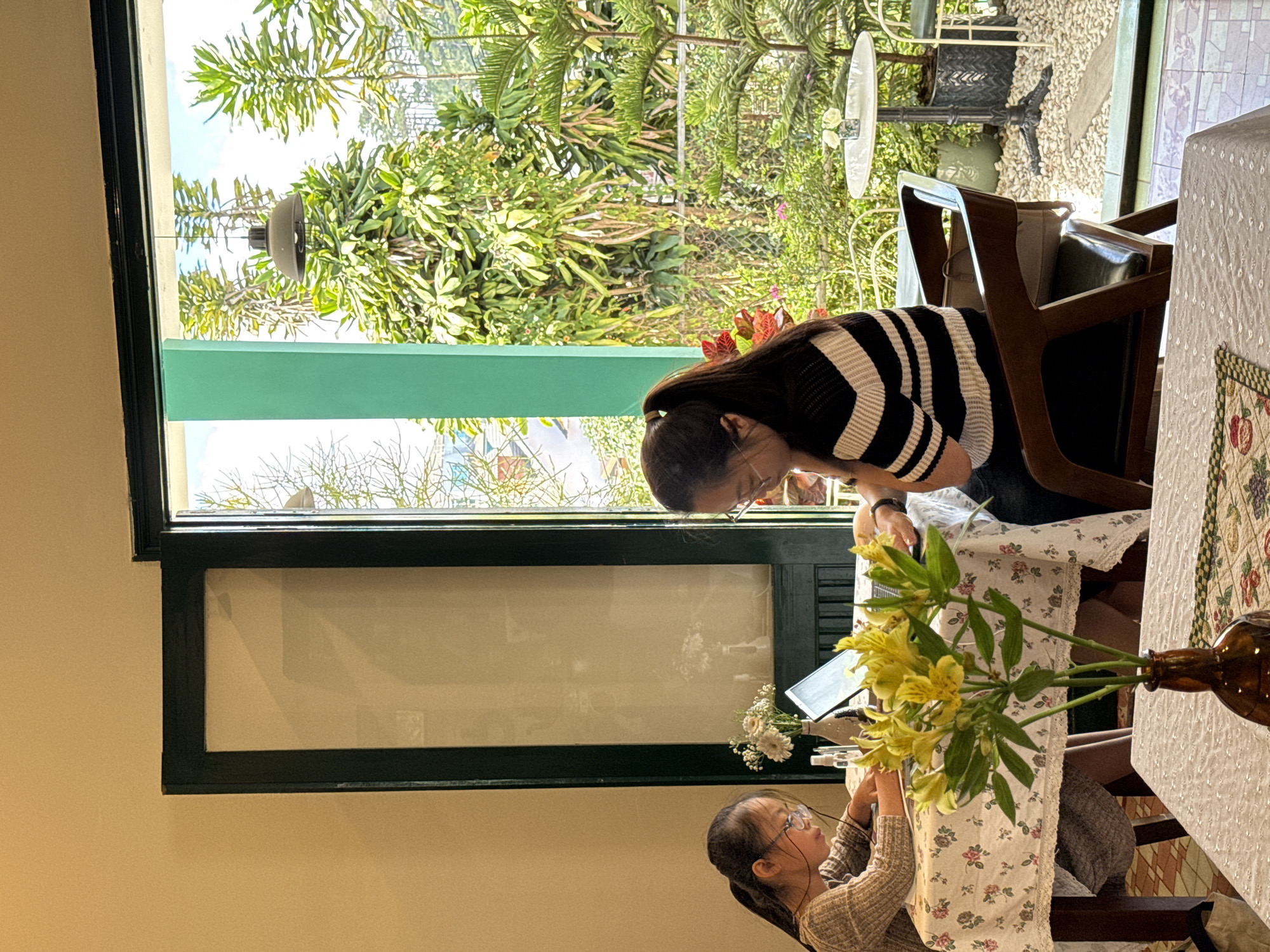 The image shows an indoor scene at a table in what appears to be a restaurant or cafe in Da Lat, Vietnam. Two people are present. One person, wearing a black and white striped shirt, is seated at the table and is looking down and to the left. Another person, wearing a tan sweater, is standing or sitting at the table and using an electronic device. On the table is a brown glass bottle with yellow flowers and leaves. There is a window in the background with green trim. Through the window, dense green foliage is visible, including trees and other plants. A white table and chairs are visible in the background through the window. The scene is illuminated by natural light.