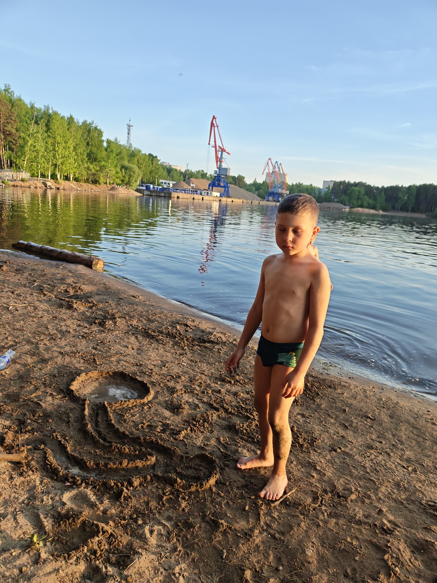 A young boy stands on a sandy beach next to a body of water. He is wearing dark-colored swimming trunks. A drawing in the sand is next to him, resembling a mushroom or an abstract design. In the background, across the water, are trees, docks, industrial cranes, and buildings. The scene is likely taken in the city of Chaikovsky, Russia, given the industrial infrastructure near a body of water.