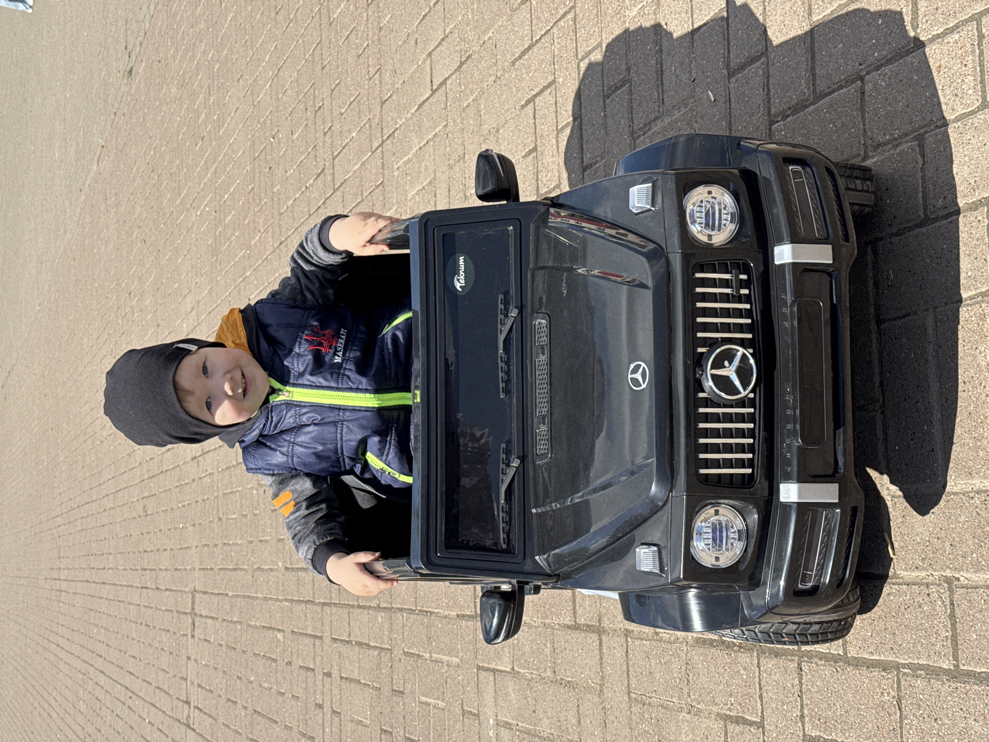 A young boy wearing a dark gray hat and a navy blue jacket with yellow trim sits in a black toy Mercedes-Benz G-Class SUV on a paved surface. The SUV is positioned at an angle in the frame, with the boy visible behind the windshield, holding onto the top of the door frame. The environment appears to be an outdoor area paved with light brown bricks. The location is Zav'yalovo, Russia. A shadow cast by the SUV and the boy is visible on the pavement.
