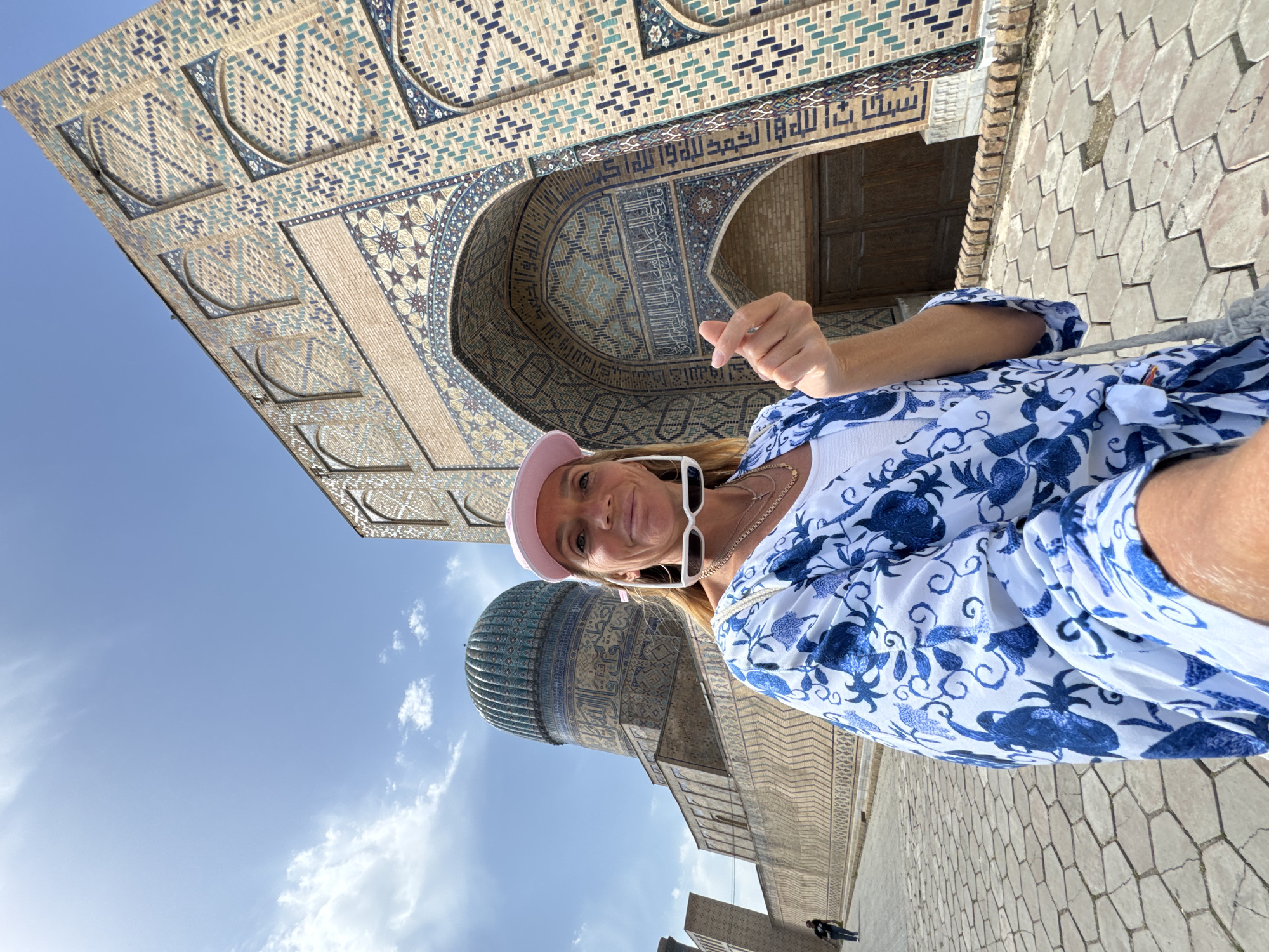 A medium eye-level shot features a woman in front of the Bibi Khanym Mosque in Samarkand City, Uzbekistan. She wears a white top, a blue and white floral blouse, a pink hat, and sunglasses. She is holding her hand in a pinch gesture, smiling, and looking into the camera. The mosque is adorned with intricate blue and white tile work. The sky is light blue with some clouds. In the distance, there are two people walking away from the camera. The ground is covered with light-colored stone tiles.