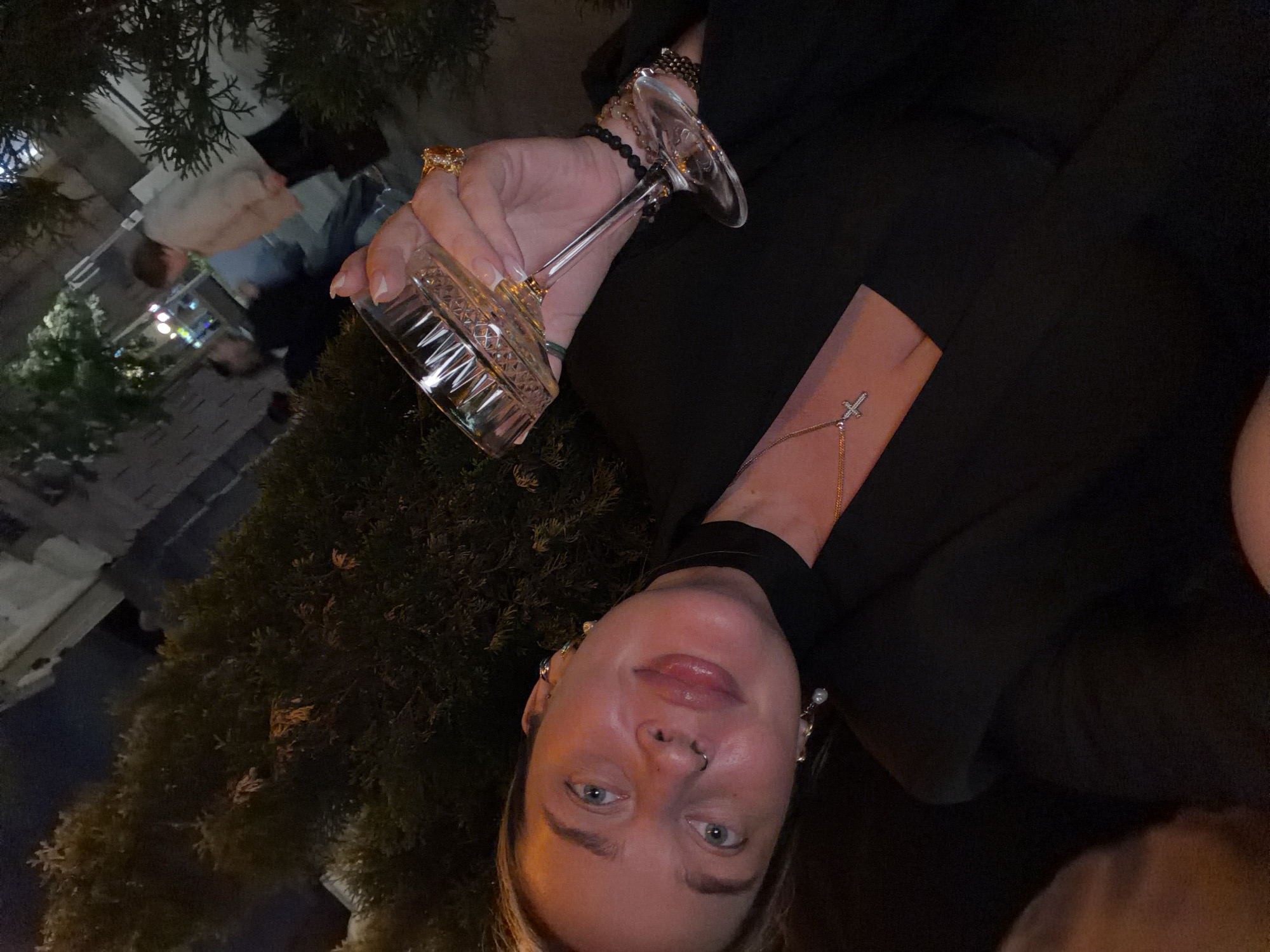 A woman with blue eyes and a nose piercing holds an empty crystal cocktail glass. She is wearing a black top and a thin gold chain bracelet with a small cross charm. She has multiple earrings in both ears. She wears an ornate gold ring on her right hand. The background includes a green shrub, other patrons, and building lights. The scene is possibly outdoors at night in a dimly lit urban setting. Given the stylistic elements and ambiance, it is not possible to confirm the location is Moscow, Russia from this image alone.