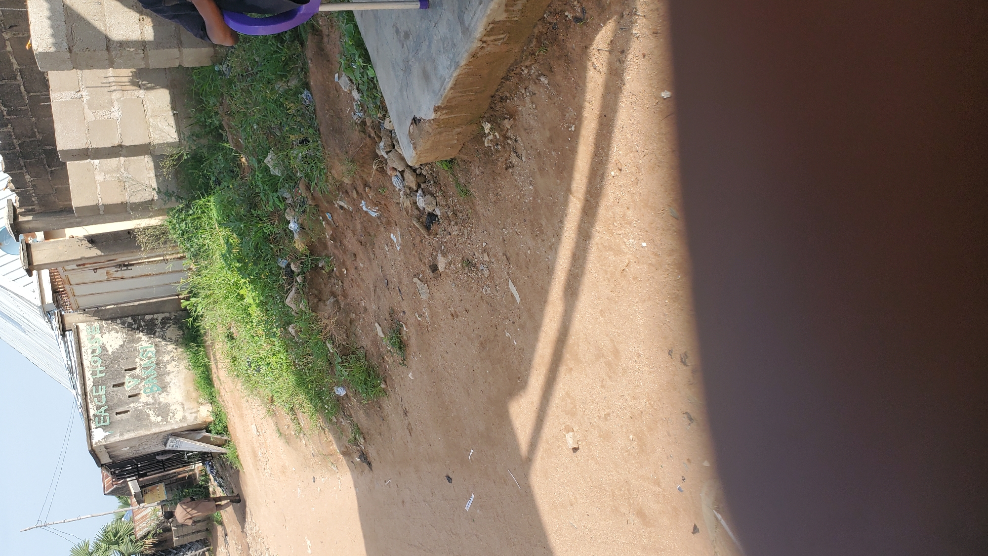 The image depicts an outdoor scene in Jalingo, Nigeria, with a focus on a dirt road leading past some buildings and vegetation. In the background, a man in a brown tunic is walking away from the camera on the left side of the road. To his left is a building with faded writing that appears to say "PEACE HOUSE" and "ABANASI". The building is weathered and shows signs of disrepair, with patches of peeling paint. Adjacent to this building is a metal gate. Further to the right, a wall constructed of concrete blocks forms a boundary. In the foreground, the ground is a mix of sand and scattered debris, including small rocks and bits of paper. A portion of a concrete structure, possibly a step or platform, is visible on the right side, with a purple chair partially visible on it. Shadows from unseen objects cast long, diagonal lines across the dusty ground. The environment appears to be a typical urban or semi-urban setting in Nigeria, with elements of local architecture and street life.