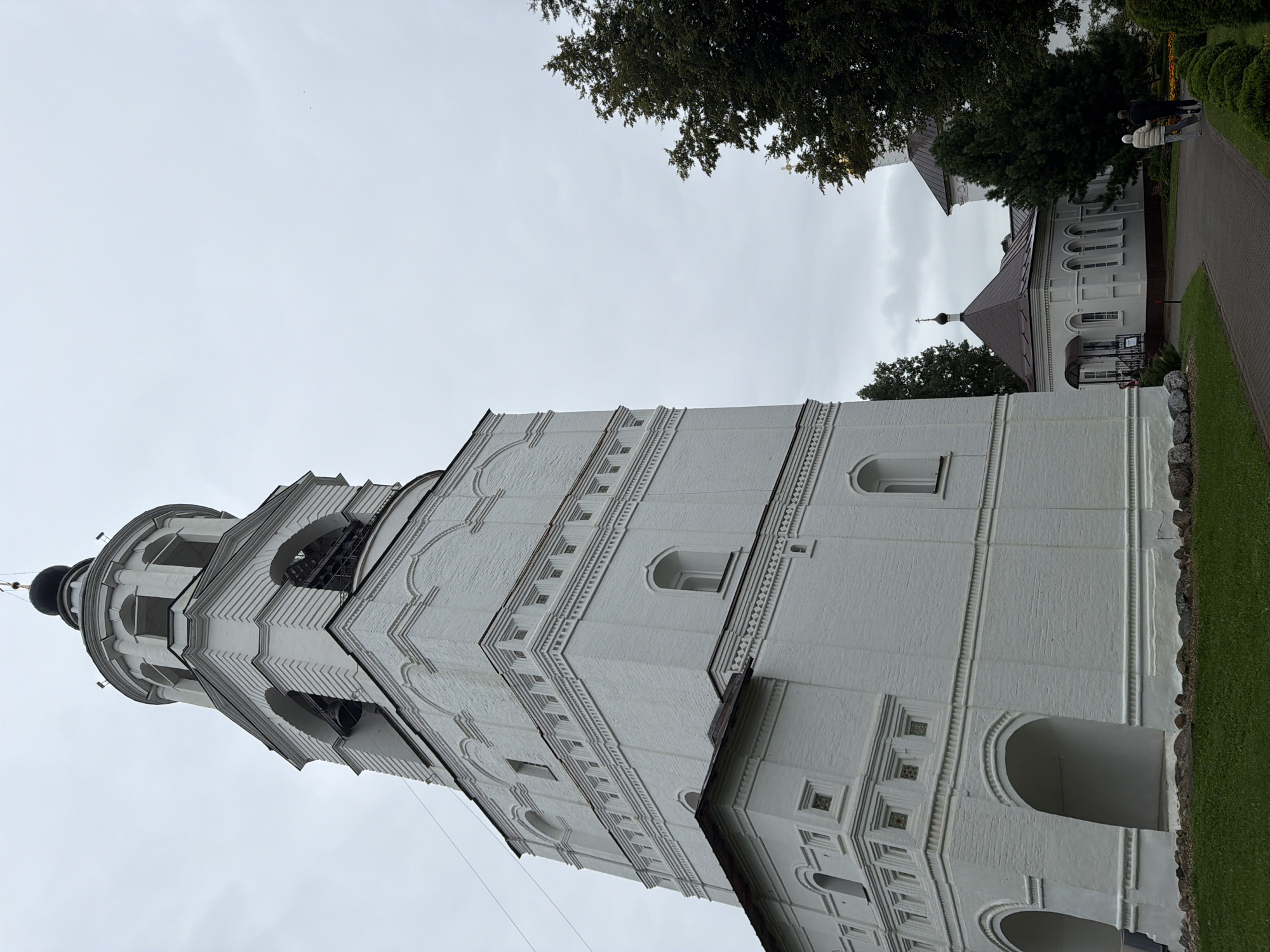 In Rusanovo, Russia, a tall, white brick bell tower dominates the left side of the frame under a cloudy sky. The tower features multiple tiers, arched openings revealing bells on an upper level, and decorative brickwork. To the right of the bell tower, a smaller white building with a brown roof and arched windows is visible, with two figures walking away from the viewer on a paved pathway. Dark green trees flank the pathway and the buildings, with a patch of green grass in the foreground.