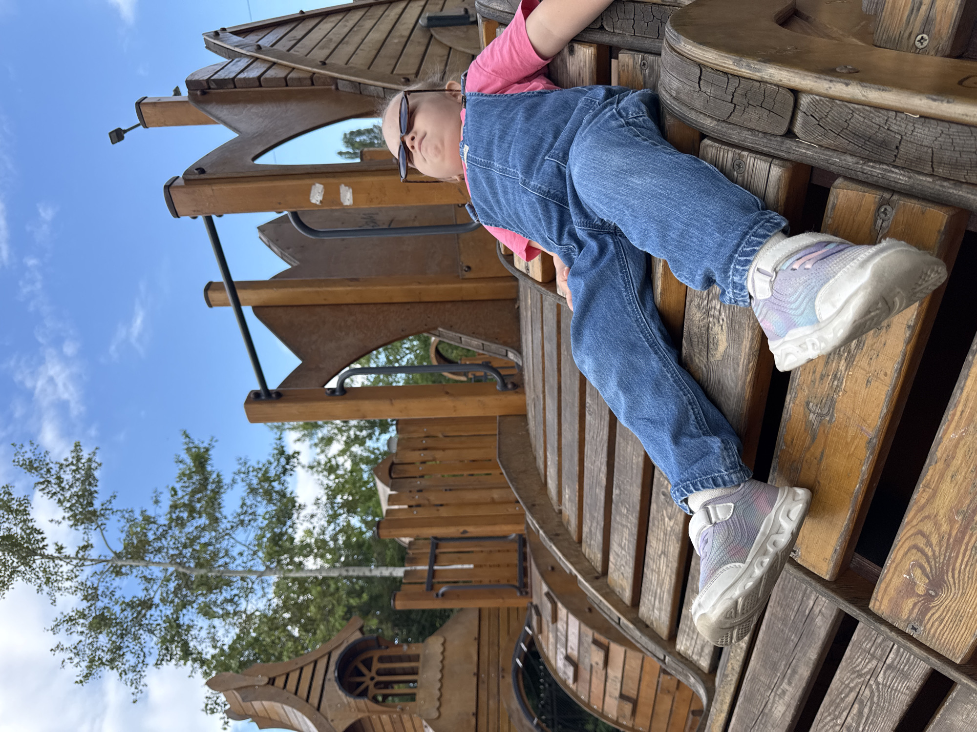 In this vertically oriented medium shot, a young child is lying down on a wooden playground structure. The child, wearing blue denim overalls over a pink long-sleeved shirt, has their legs extended and is looking upwards, away from the camera. They are wearing dark sunglasses. The playground appears to be made of weathered wood and has various levels and wooden planks. In the background, the sky is a clear blue with some scattered white clouds. Green trees with sparse foliage are visible, suggesting a natural outdoor setting. The overall lighting and cloud cover indicate it is daytime, likely a sunny day. The child's relaxed pose and upward gaze might suggest a moment of rest or observation. The image seems to be taken in a park or a playground area. There is no visible text in the image. The location context provided is Верхнерусское, Russia.