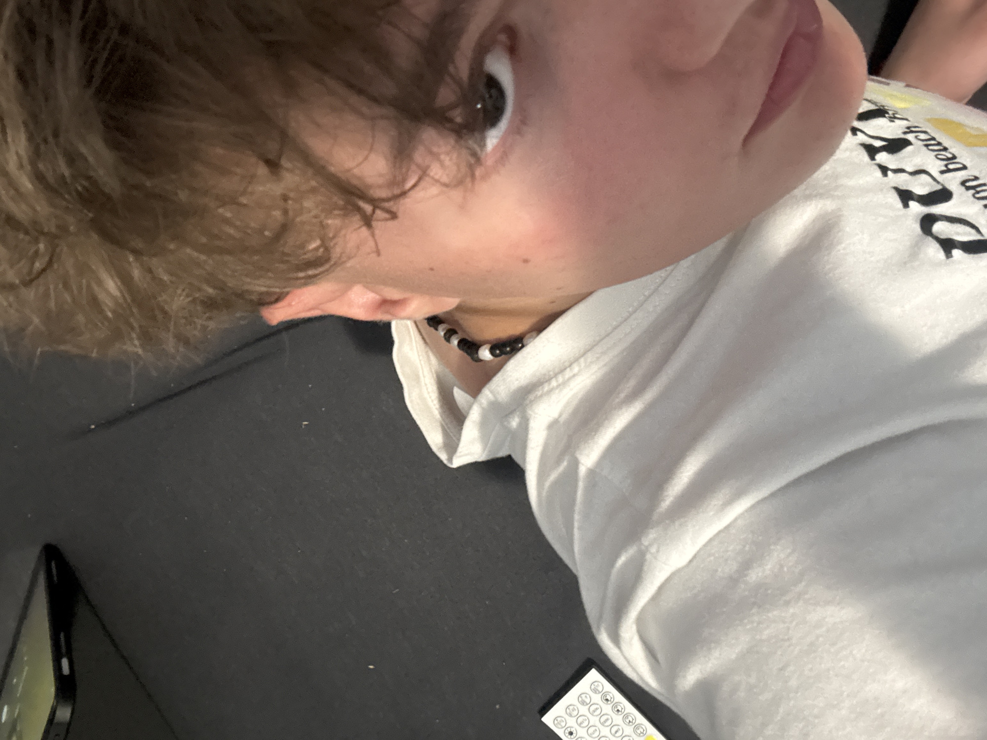 The image is a close-up, angled selfie of a person with light brown hair, fair skin, and an eye visible in the upper left. The person is wearing a white t-shirt with black lettering, partially obscured by the angle, and a black and white beaded necklace. A black smartphone is visible in the lower left corner of the frame. A white remote control with several buttons is located near the lower right corner. The background appears to be a dark, possibly black, surface. There is no information to indicate the location is in Troyan, Bulgaria.