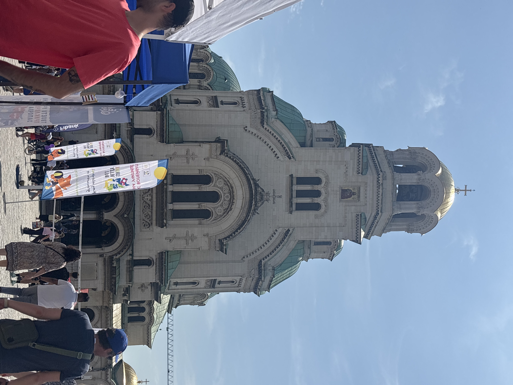 The image shows a view of the Alexander Nevsky Cathedral in Sofia, Bulgaria. The cathedral is a large, ornate building with multiple domes, partially obstructed in the lower left corner by other golden domes. The sky is clear and blue. People can be seen in the foreground, including individuals wearing t-shirts and one wearing a backpack. There are flags and banners with colorful designs and Cyrillic text set up on the plaza in front of the cathedral. Part of a tent or awning is visible in the upper part of the frame, as well as the upper body of a person in a red shirt. The image appears to be taken at ground level from a public square.