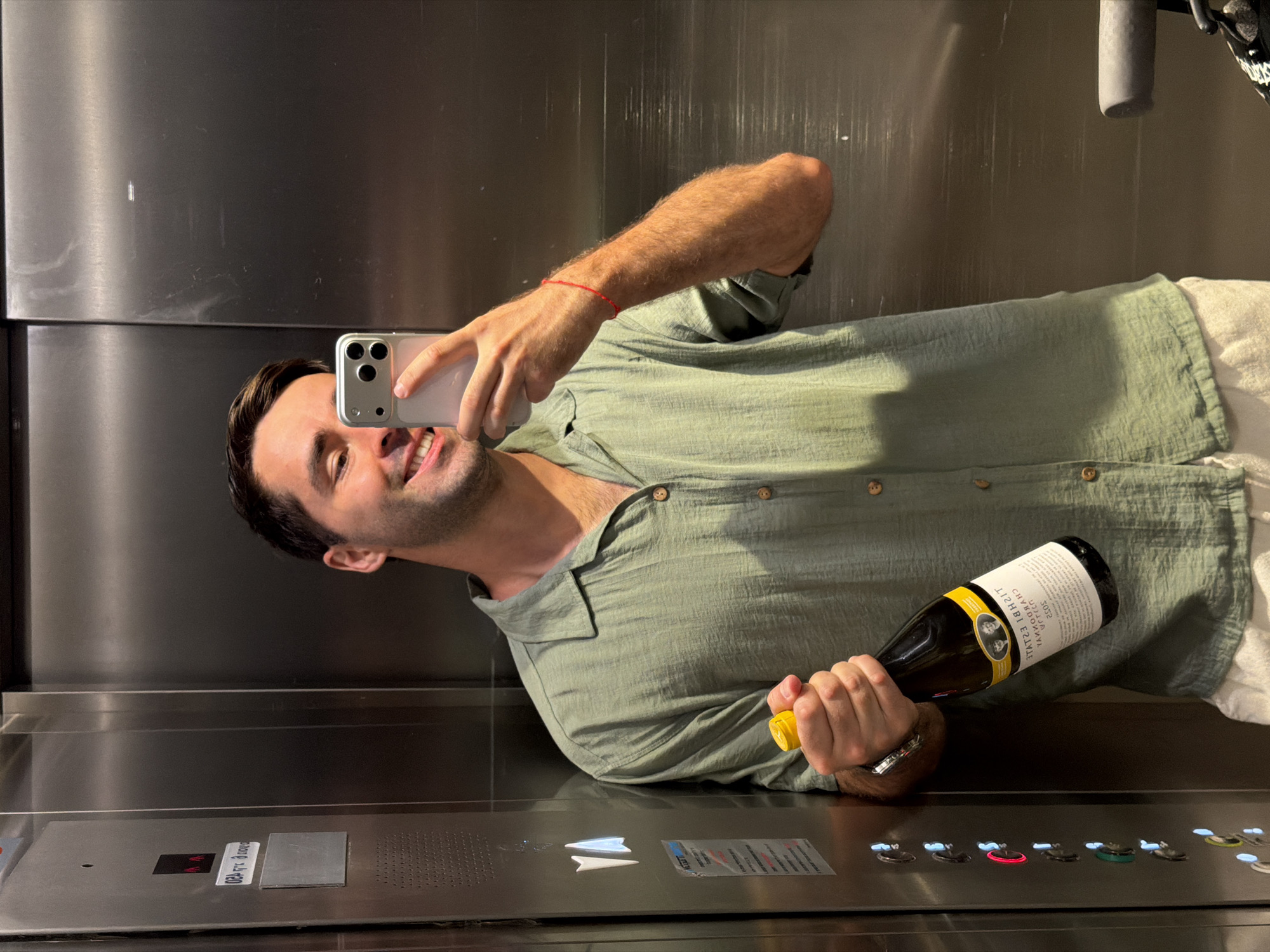 A man is taking a selfie in an elevator. He is holding a bottle of wine in his left hand and a smartphone in his right. The man is wearing a green collared shirt and a red bracelet. The elevator appears to be made of stainless steel with an illuminated control panel. The control panel has an inscription that seems to be in Hebrew. Although the location can't be conclusively determined, the presence of Hebrew inscriptions suggests it's likely in Israel, possibly Tel-Aviv as requested.