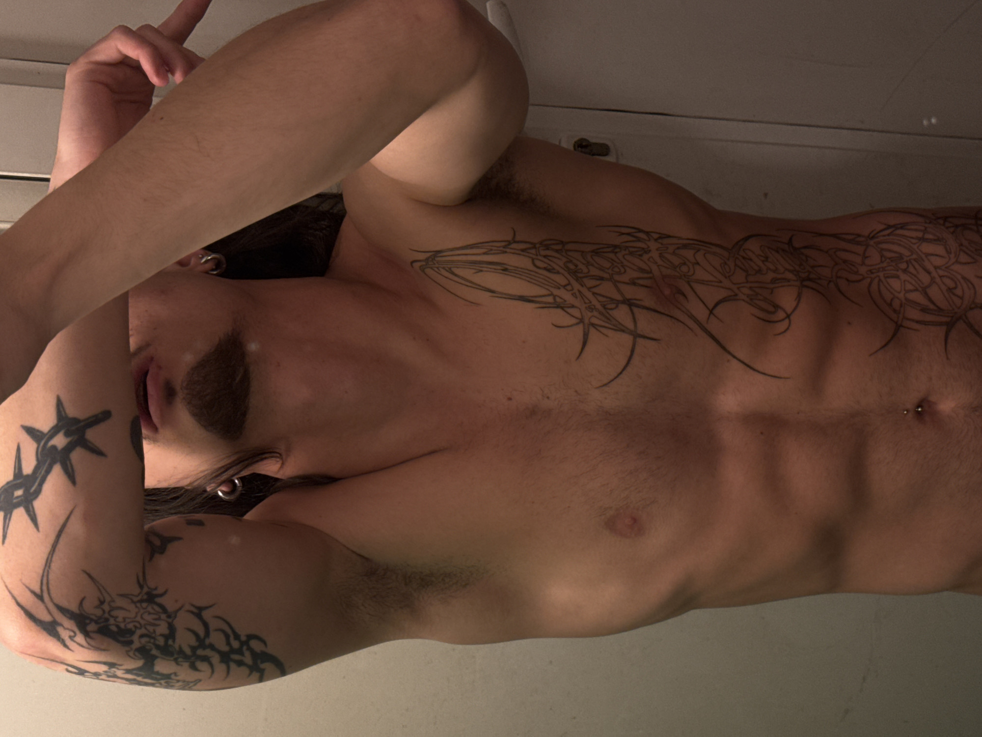 The image is a close-up, waist-up shot of a shirtless individual, likely male, posing for a mirror selfie. The person has several tattoos: a large tribal-style tattoo on the left arm and shoulder, and a long, intricate tattoo running down the right side of their torso, from the chest to the abdomen. The individual has a goatee and is wearing a silver hoop earring in the left ear. They also have a small silver belly button piercing. The background appears to be an indoor setting, possibly a bathroom, with a plain wall and a door visible on the right. The lighting is somewhat dim, casting shadows and highlighting the musculature of the person's abdomen and arms. The overall impression is one of self-presentation. There is no information in the image to suggest a location in Sofia, Bulgaria.