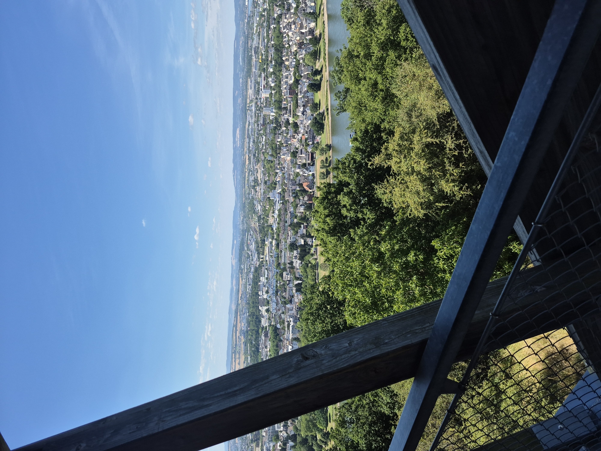 The image shows a panoramic view of Koblenz, Germany, from a high vantage point. In the foreground, part of a wooden observation deck or tower is visible, with metal railings and wire mesh fencing. Beyond the deck, lush green trees dominate the middle ground, with a wide river, likely the Rhine, flowing through the city. The far background reveals the sprawling urban landscape of Koblenz, characterized by a mix of residential buildings, commercial structures, and industrial areas, all set against a backdrop of rolling hills and a clear blue sky with scattered white clouds. The sun appears to be shining brightly, indicating a daytime scene, likely during the summer months due to the full foliage of the trees. There are no people visible in the image.