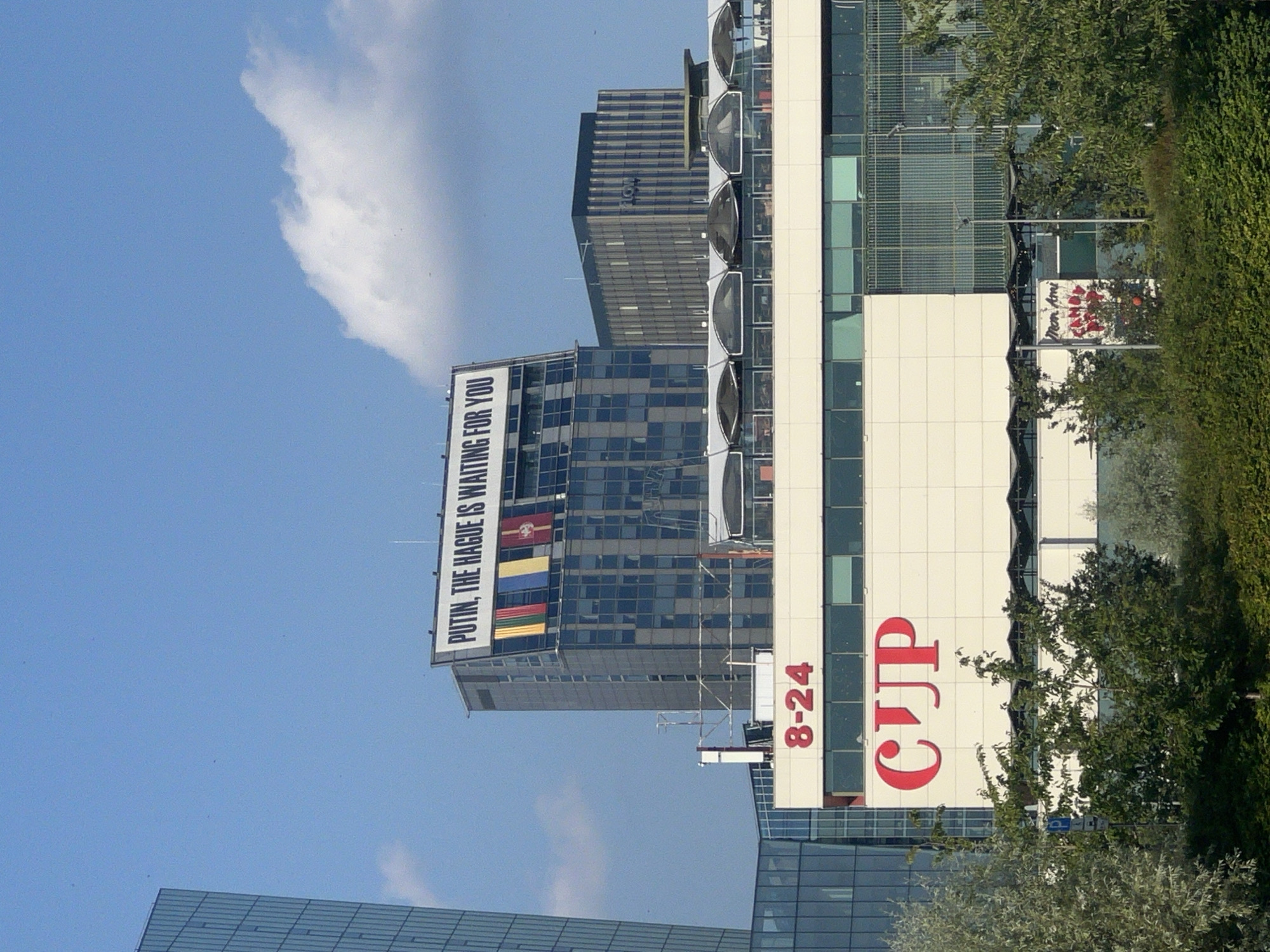 The image shows a cityscape with several buildings under a bright blue sky with some clouds. The most prominent building, a tall, dark-colored skyscraper, features a large billboard at its top that reads "PUTIN, THE HAGUE IS WAITING FOR YOU" in white letters against a black background. Below the billboard, several flags are displayed, including what appear to be the flags of Ukraine and other European nations.
In the foreground, a lighter-colored, modern building has a red sign with "8-24" in large numbers and another red sign with the stylized letters "CUP". To the right of the "CUP" sign, a smaller white sign with red lettering says "Mon Ami" and "CANDY SHOP". The buildings are surrounded by lush green trees and foliage, suggesting an outdoor urban setting. The overall impression is of a modern city, likely in Europe, given the political message on the billboard and the style of architecture.
The location is identified as Vilnius, Lithuania.
