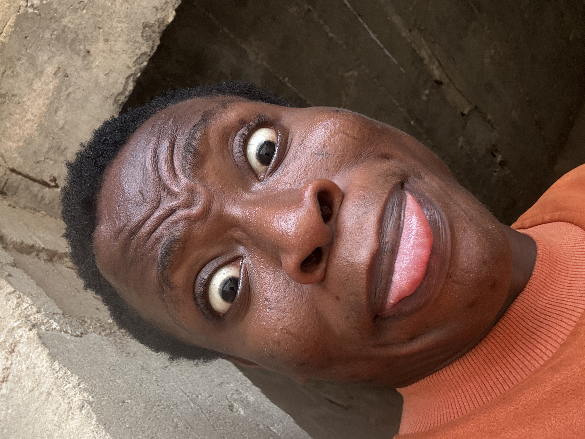 A close-up, slightly angled selfie of a young Black man. He is sticking his tongue out slightly, and his eyes are wide. He has short, dark hair and is wearing an orange shirt. The background is a combination of concrete and shadow, possibly indicating he's in or near a structure. The location is Unknown, Unknown.