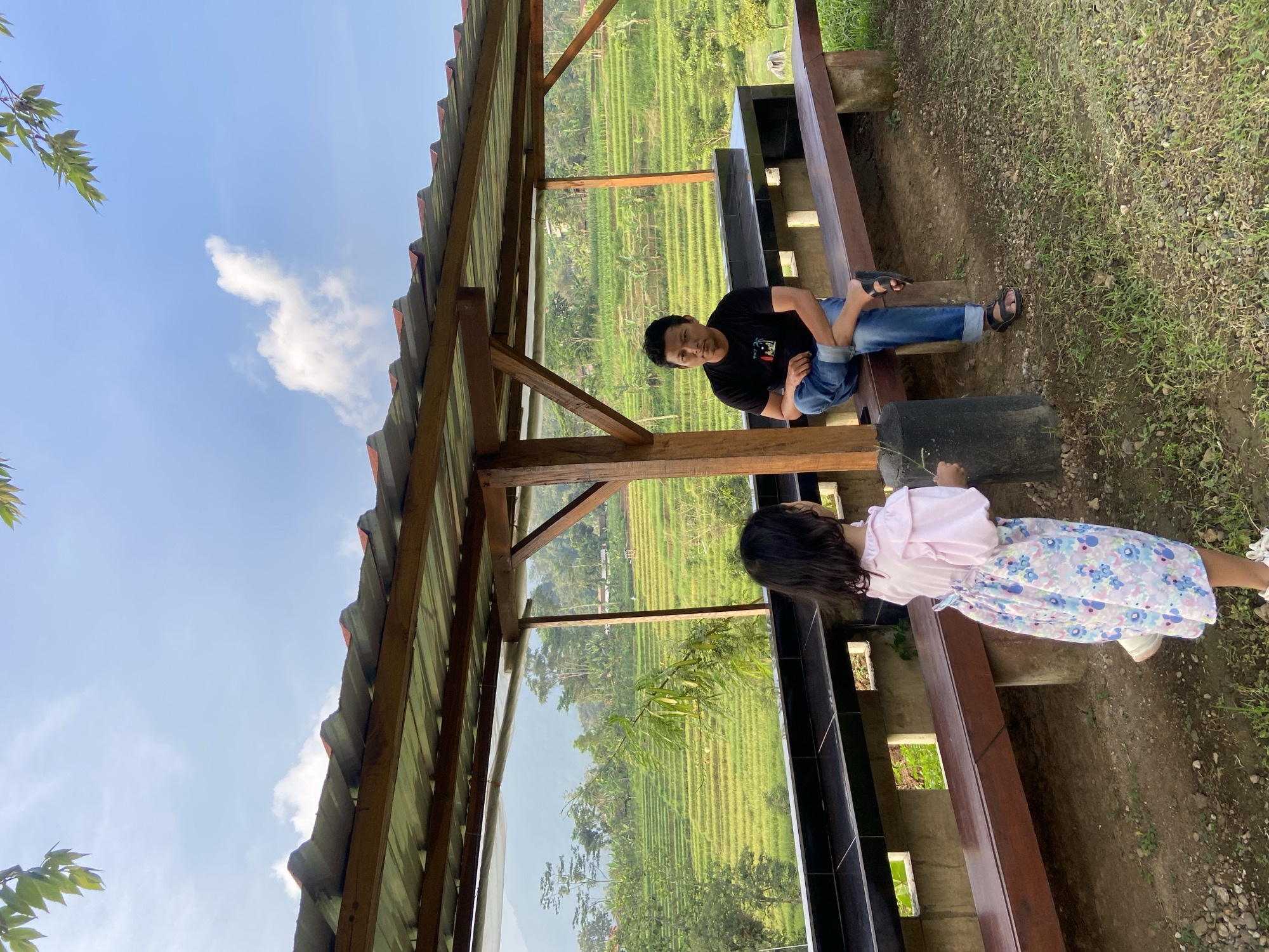 Two individuals are situated within a wooden structure, possibly a gazebo or shelter. The structure is in Penanggungan, Indonesia. The person further from the camera is an adult male. He is seated on a wooden bench, wearing a black t-shirt and jeans. His dark hair is short. The other person is closer to the camera and appears to be a girl, wearing a pink top and a light blue floral skirt. She is standing near the edge of the bench, seemingly looking out. Behind the structure, a view of terraced fields and vegetation can be seen. The sky is blue with some clouds. The ground outside the structure is covered with grass and gravel.