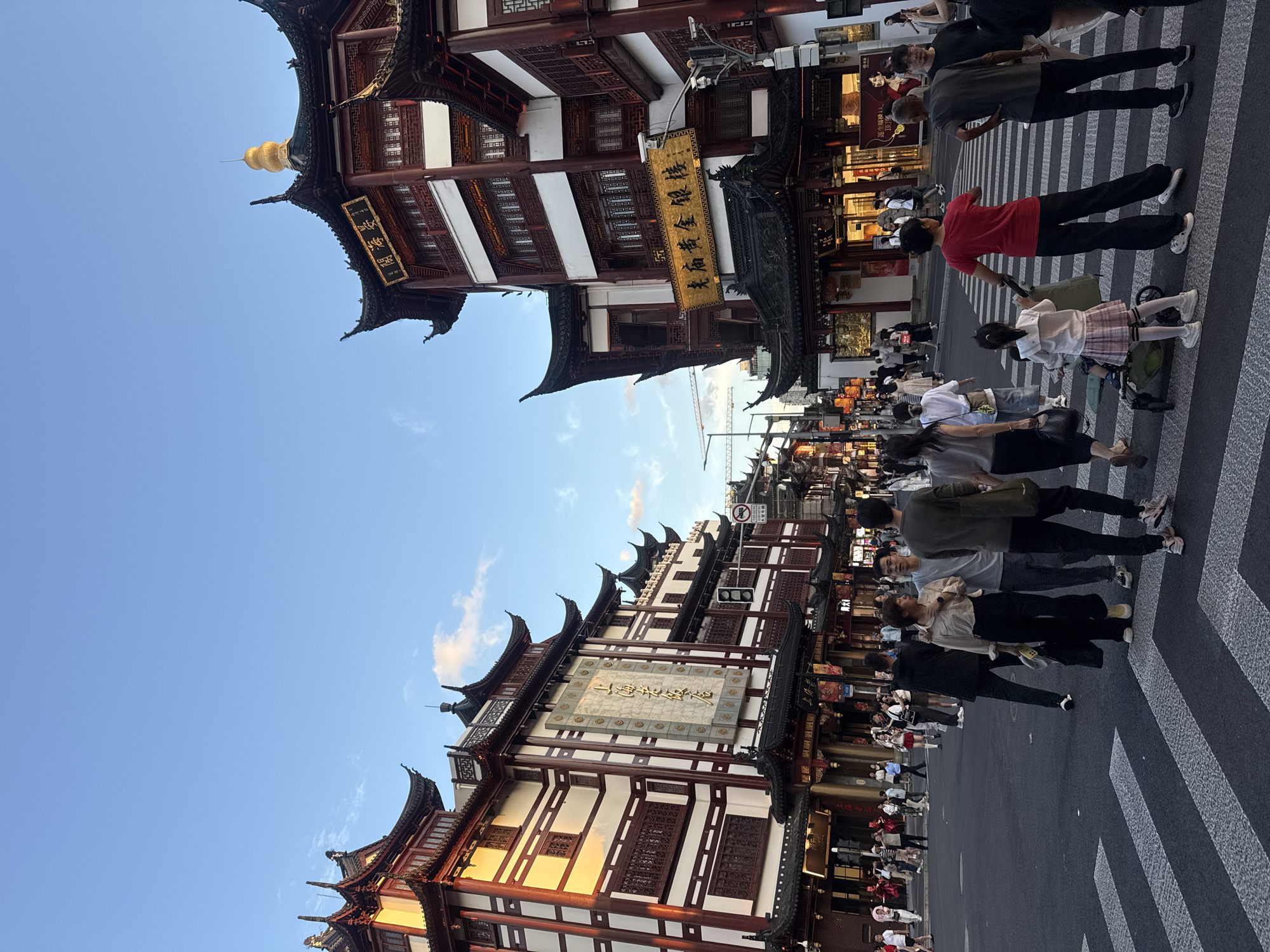 The image depicts a street scene in Shanghai, China, likely within a historic district such as Yu Garden or a similar area characterized by traditional Chinese architecture. In the foreground, a crosswalk is visible with several pedestrians crossing. Among them are two individuals, a man and a woman, walking arm-in-arm, and a woman pushing a stroller with a child. Further back, more people are walking and interacting.
The buildings on either side of the street are a dominant feature, exhibiting traditional East Asian architectural styles. They are multi-story structures with prominent curved roofs, dark wood accents, and white or light-colored walls adorned with red detailing. Several signs with Chinese characters are displayed on the buildings. The sky above is a clear blue with a few wispy clouds, suggesting it is either early morning or late afternoon, as indicated by the warm light on the buildings. The overall impression is a bustling urban environment with a strong cultural and historical presence.