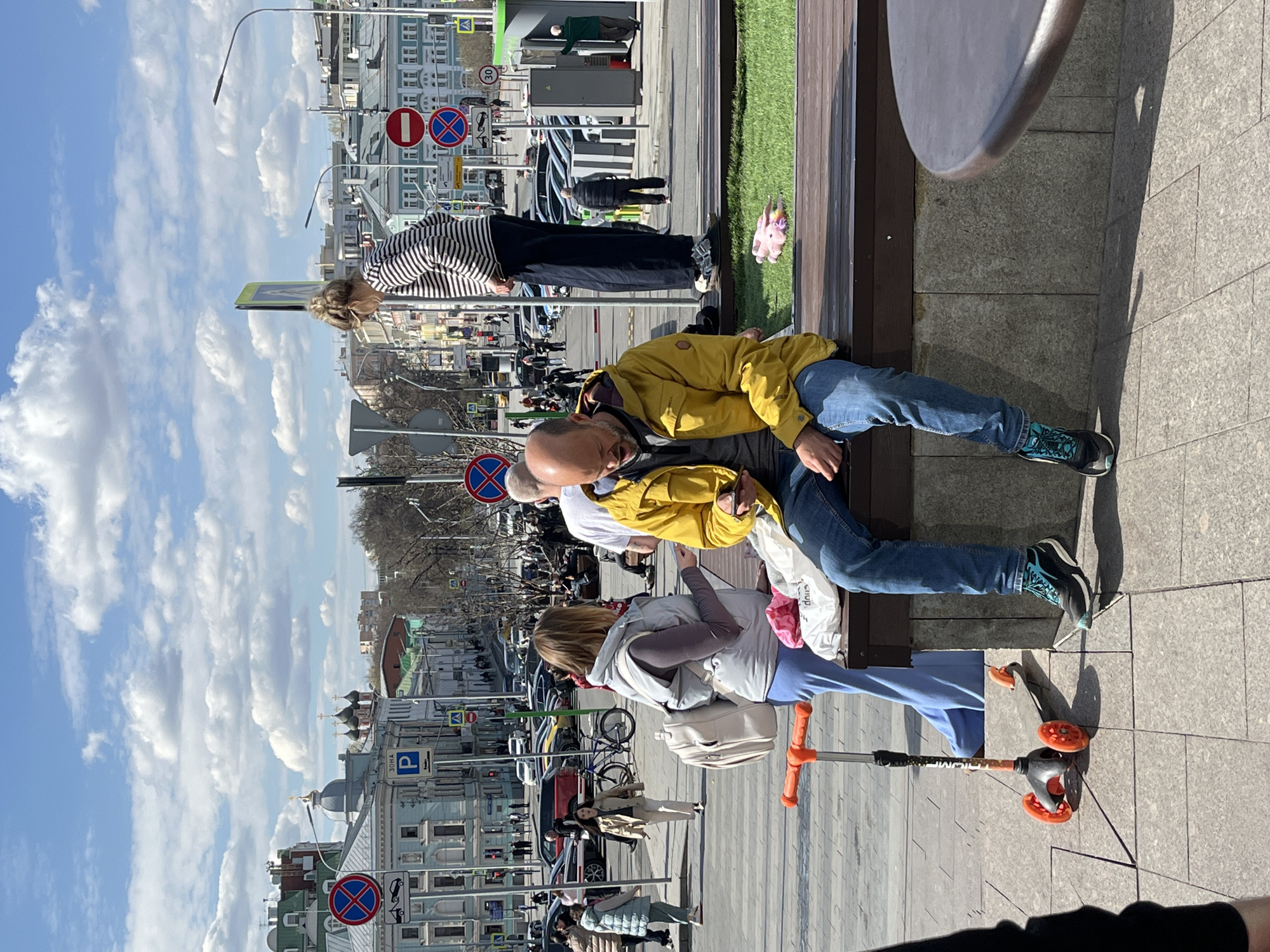The image shows a street scene in Moscow, Russia. A man in a yellow jacket and jeans is sitting on a bench. Next to him, a woman in a white jacket and blue pants is standing with a scooter. Another person is standing nearby in a black and white striped shirt and black pants. Other people are walking in the background, and cars are driving on the street. Traffic signs and buildings are visible in the background, and the sky is blue with clouds.