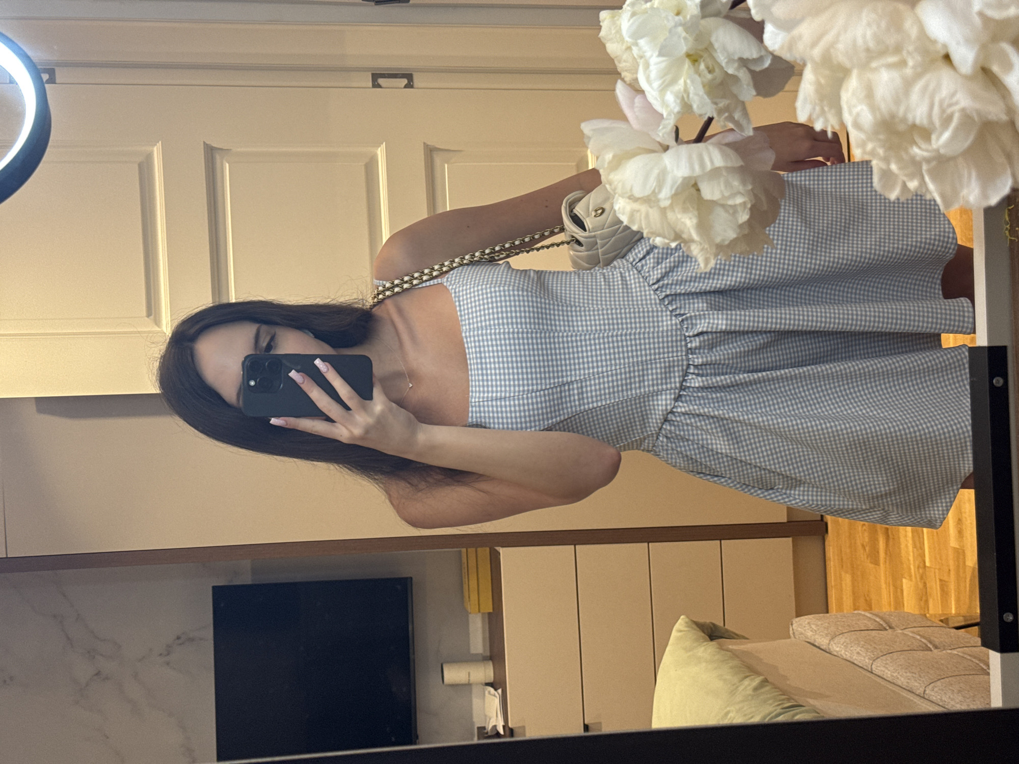 A mirror selfie captures a person, likely female, wearing a light blue and white gingham-patterned strapless dress with a ruffled bodice and a full skirt. The person holds a black smartphone, obscuring their face, and their long, dark hair falls over one shoulder. They accessorize with a small, light-colored quilted handbag featuring a gold chain strap draped over their arm, and a delicate necklace. Their fingernails are long and appear to have a pale manicure.
The reflection in the mirror reveals an indoor setting, possibly a bedroom or dressing area. To the left, a white paneled door with dark hardware and a bright ring light are visible. On the right, a large arrangement of white, artificial flowers (resembling peonies) partially frames the scene. Below the door, a dark rectangular screen (possibly a television) is set into a light-colored cabinet or wall unit. Further right, part of a bed with a light-colored, possibly tufted headboard and a pale green pillow can be seen. The floor in this area is wooden. Additional background elements include light-colored cabinets, shelves holding a yellow object (perhaps a book), and a white dispenser with a folded cloth. The scene is brightly lit, suggesting an indoor environment during the day or with artificial lighting. There is no visible text.