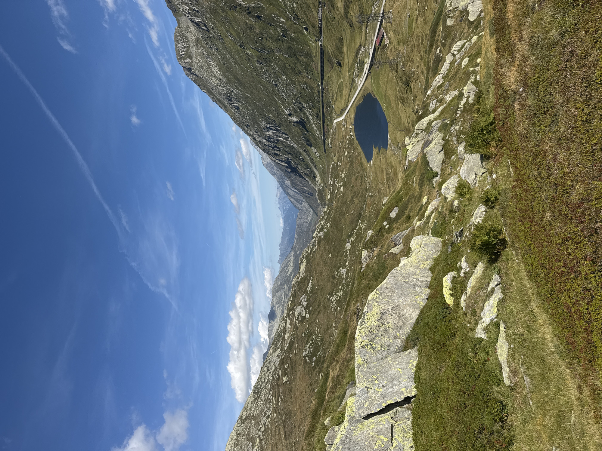 The image depicts a mountainous landscape near Airolo, Switzerland, under a clear blue sky with scattered white clouds and contrails. The foreground is dominated by rocky terrain covered in low-lying vegetation, with large, lichen-covered boulders. In the mid-ground, a valley opens up, featuring a dark, heart-shaped lake and a road winding alongside it. A train track runs parallel to the road, with a dark tunnel entrance visible on the mountainside. The right side of the image is characterized by a steep, grassy mountain slope, with some visible electrical pylons and a red-roofed building at its base. The background shows a range of distant, hazy mountains. There are no people or animals visible in the scene.