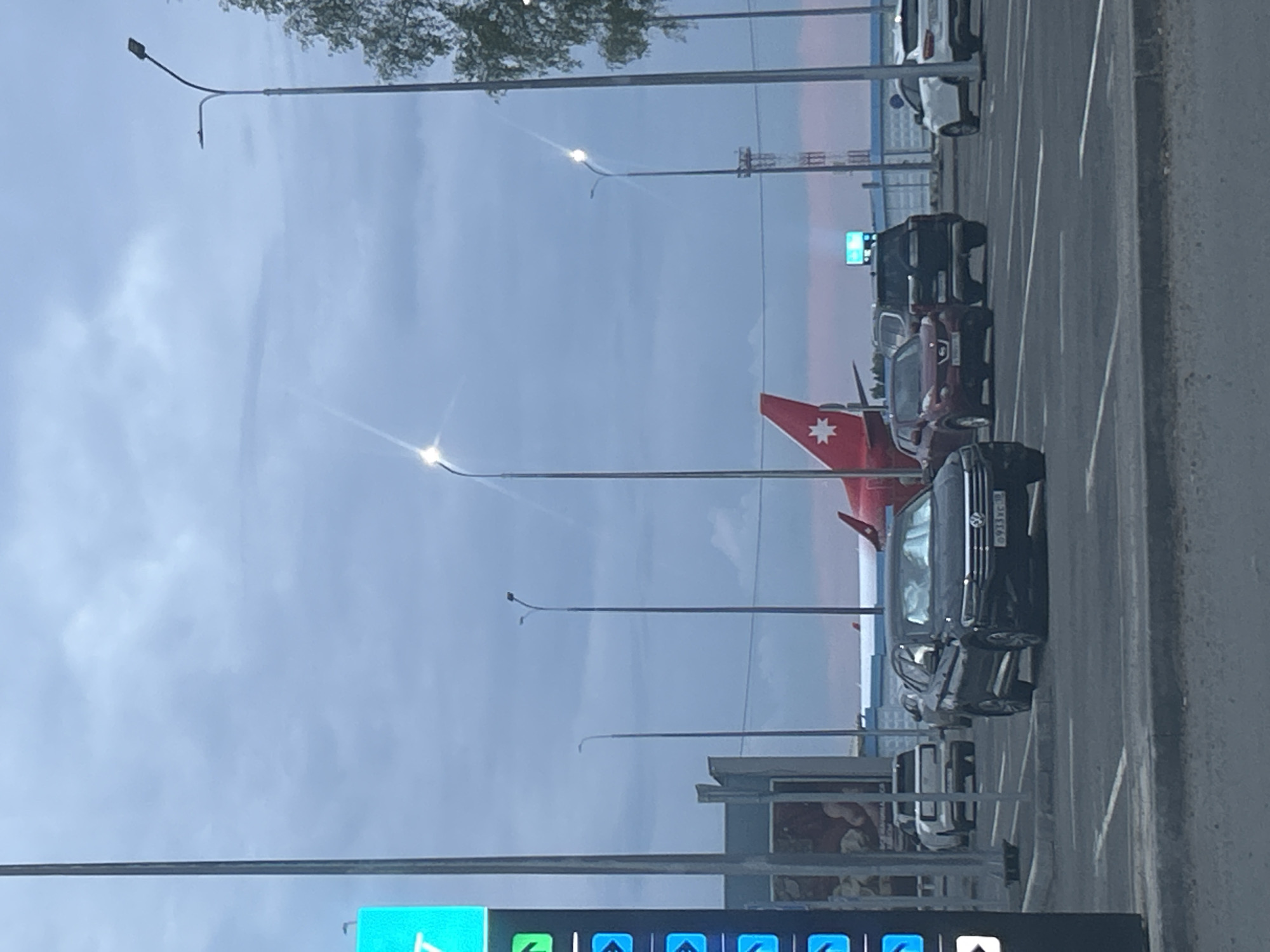 The image shows a parking lot with several cars and an airplane in the background. A red airplane with a white star logo on its tail is visible, with the fuselage extending to the right. Multiple vehicles are parked in the lot, including a black Volkswagen sedan in the foreground with a Russian license plate that reads "0933XC 77". Other cars include a white SUV, a dark SUV, and a red compact SUV. Several streetlights are present, with one in the foreground illuminating a portion of the parking lot. A tall pole with a blue digital sign is also visible to the left. The sky is overcast, suggesting a cloudy day. The scene appears to be at an airport or a related facility.