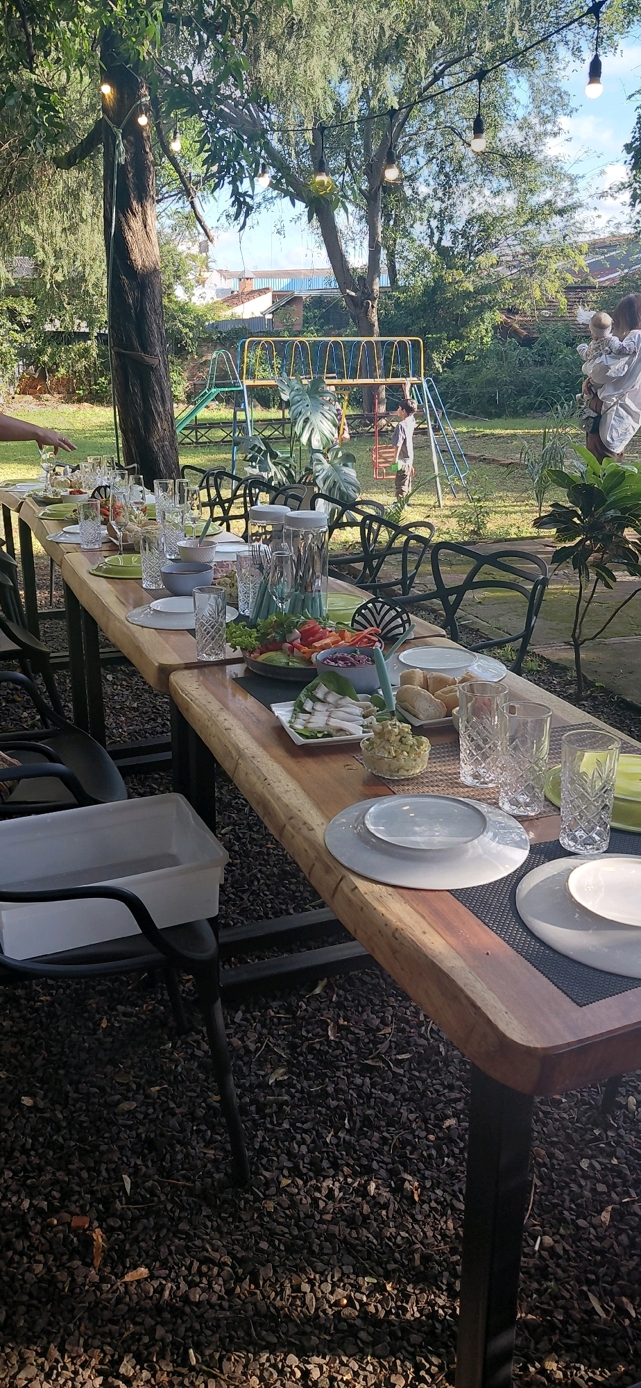 A long, wooden table is set for a meal outdoors in a garden setting, likely in Encarnación, Paraguay, given the presence of string lights and a playground in the background. The table is laden with dishes, glasses, and cutlery, suggesting a gathering. In the background, a playground structure with colorful bars is visible, and a person is holding a baby, while another person stands near the playground. The ground is covered in dark gravel, and lush green trees provide shade over the scene.