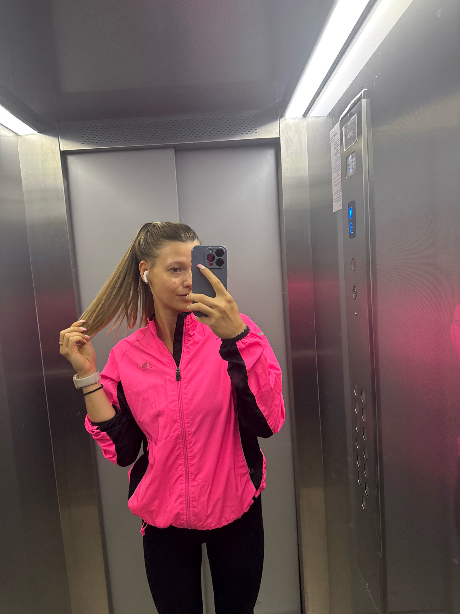 A young woman with blonde hair pulled into a high ponytail is taking a mirror selfie in an elevator. She is wearing a bright pink athletic jacket with black accents, black leggings, wireless earbuds, and a smartwatch. Her right hand is holding her phone, which has a dark case, and her left hand is touching her ponytail. The elevator is made of brushed metal, with a bright fluorescent light panel on the ceiling. The elevator control panel is visible on the right side of the frame, showing a blue illuminated 'E' and various buttons. The overall environment suggests a modern, functional space. There are no discernible landmarks that would place this image in Sofia, Bulgaria.