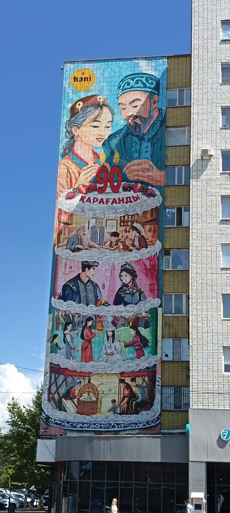 The image displays a large mural on the side of a multi-story building, depicting a sequence of scenes. The mural is predominantly illustrated with bright colors and appears to celebrate a significant anniversary, indicated by the large number "90" prominently featured on what resembles a cake. The text "ҚАРАҒАНДЫ" (Qarağandı) is visible beneath the "90," suggesting the mural is located in Qarağandı city, Kazakhstan.
The top section of the mural features a portrait of a man and a woman, likely representing a couple, against a blue sky backdrop. Below them, several vignettes unfold, showcasing different aspects of life and tradition. One scene depicts a family gathered around a table, possibly engaged in an activity or a meal. Another section illustrates a wedding or a similar ceremonial event with individuals in traditional attire. Further down, a scene depicts a cradle with adults tending to it, suggesting the theme of family and lineage.
The building itself has a light-colored brick facade with numerous windows. The mural covers a significant portion of one side of the building, extending from near the top to below the middle floors. Below the mural, a street-level view shows a sidewalk, parked cars, and a few pedestrians walking. The sky is a clear blue with some scattered white clouds. The mural's artistic style is detailed and narrative, conveying a sense of history and cultural celebration.