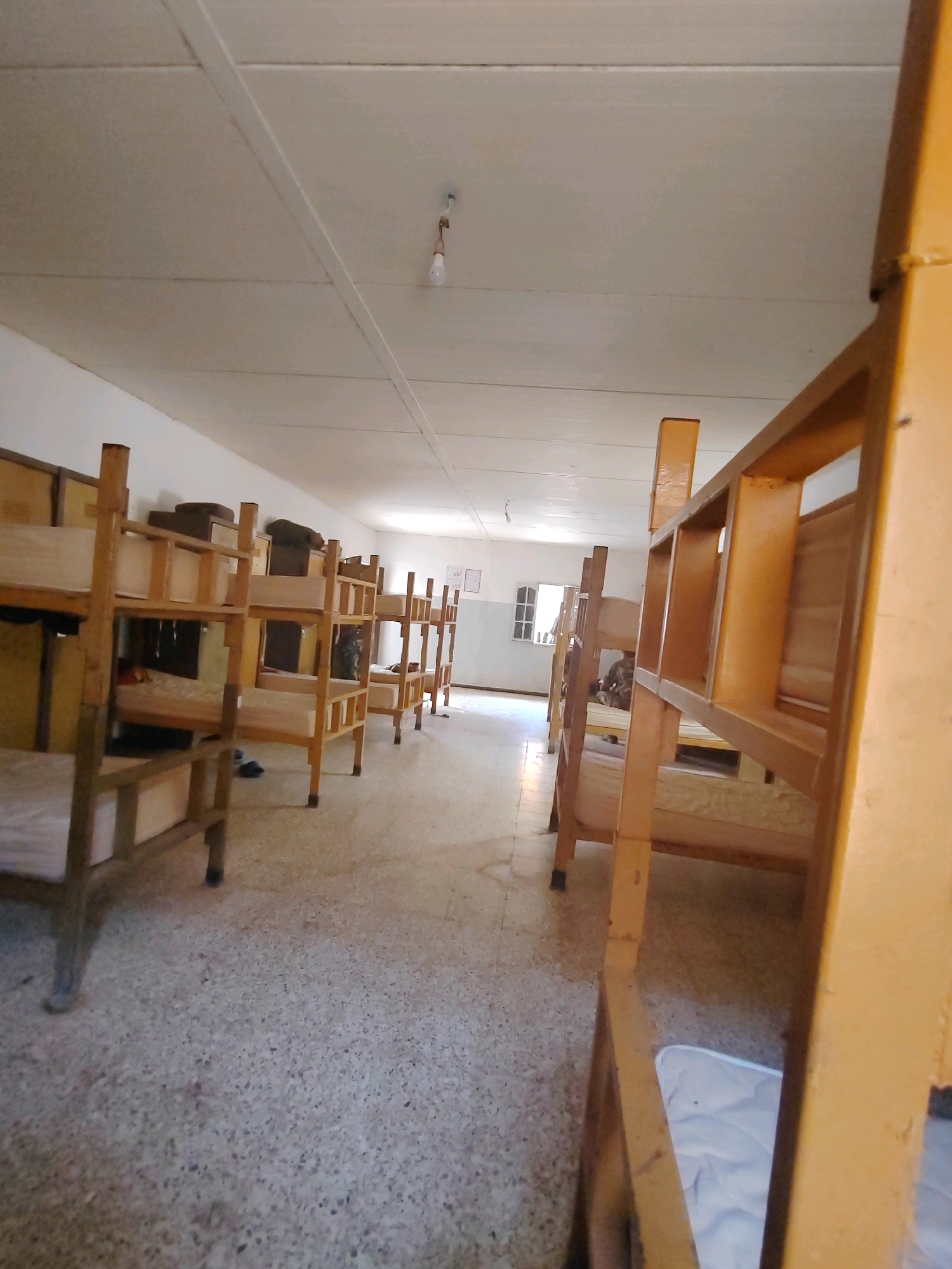 A long, narrow dormitory room in Mougheul, Algeria, is filled with rows of wooden bunk beds. The beds are simple in design, with wooden frames and mattresses. Some of the mattresses are covered with white sheets, while others appear bare. The room is lit by a single bare bulb hanging from the ceiling, and the walls are painted white. The floor is made of a speckled terrazzo material. The room appears to be empty of people, with only the beds and a few scattered personal items visible. The overall impression is one of stark utilitarianism and a lack of personal touch.