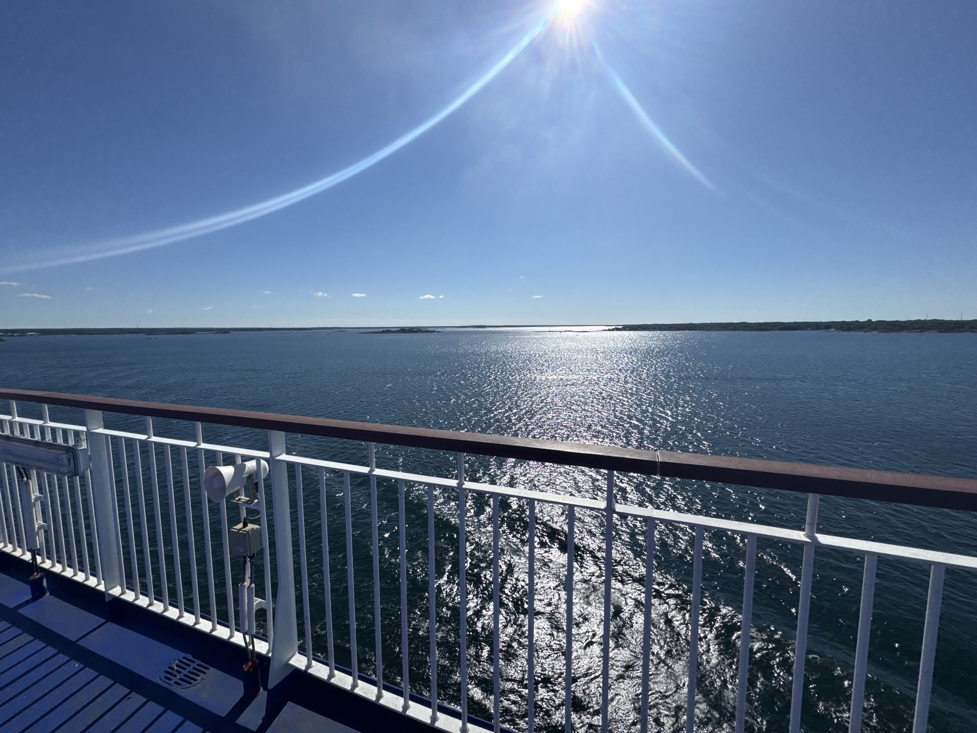 The image captures a view from a ferry deck looking out at a body of water under a clear blue sky with the sun high overhead, creating a bright glare on the water's surface. The ferry's railing, made of white metal bars with a dark brown wooden top, runs along the bottom of the frame. A speaker and some electrical equipment are mounted on the railing. The blue deck of the ferry is visible in the foreground. In the distance, a long, tree-lined coastline is visible, dotted with what appear to be small islands. The sun's rays are visible as a lens flare, and a faint, curved white contrail stretches across the sky. The location is likely the waters surrounding Karlskrona, Sweden, given the presence of coastal islands and the ferry traffic common in this archipelago.