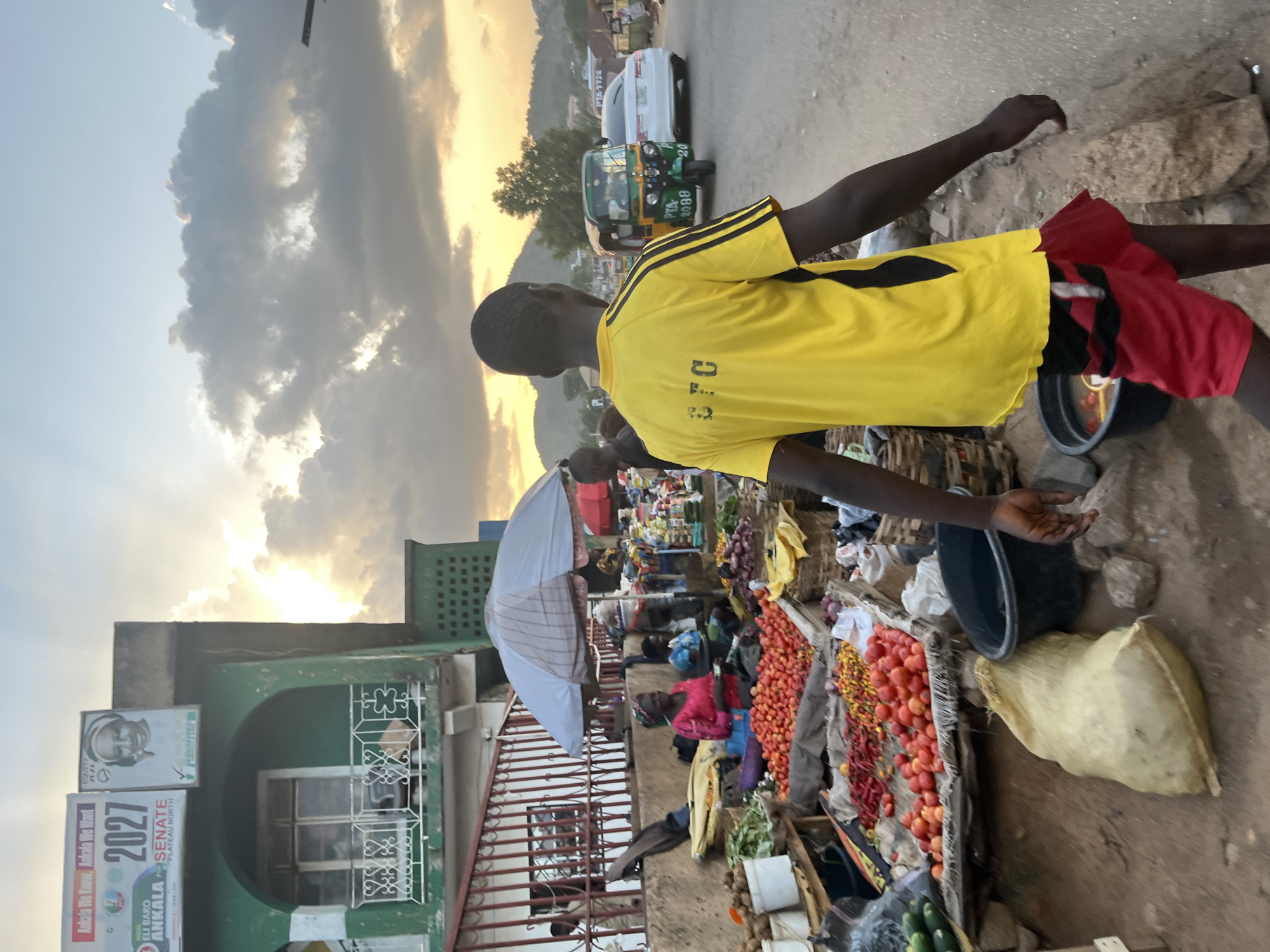A medium shot captures a bustling outdoor market scene at dusk in a city identified as Unknown, Unknown. In the foreground, a young person wearing a bright yellow t-shirt with black stripes on the sleeves and red shorts walks away from the camera, their back to the viewer. The t-shirt displays "3TC" in black lettering. To the left, market stalls are laden with a vibrant array of fresh produce, including tomatoes, chilies, cucumbers, and onions. A woman in a pink top sits behind one of the stalls, appearing to interact with customers. A blue and white striped umbrella provides shade over a stall. In the background, a green building with a balcony overlooks the market. Further down the street, a green auto-rickshaw and a white car are visible, along with the silhouettes of distant hills against a dramatic sky filled with clouds illuminated by the setting sun. The ground is a mix of dirt and pavement, with rocks and a large, light-colored sack scattered around the market area.