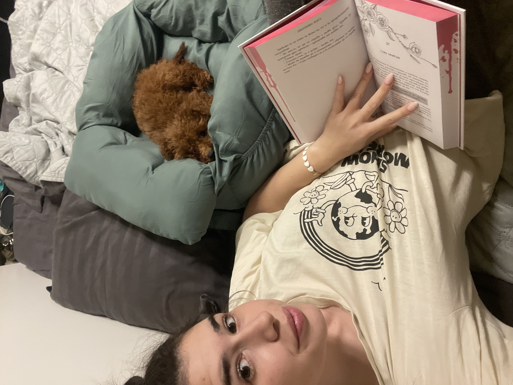 A woman is lying on a bed and reading a book with pink pages, and a small brown poodle is curled up on a green pillow next to her. The woman is wearing a light-colored t-shirt with a black cartoon design and has a white bracelet on her left wrist. She is looking at the camera. The room appears to be a bedroom, with a gray pillow and a white and gray quilted blanket visible in the background. Based on the text in the image, the book is written in Bulgarian. There's no information to determine the specific location as Sofia, Bulgaria.