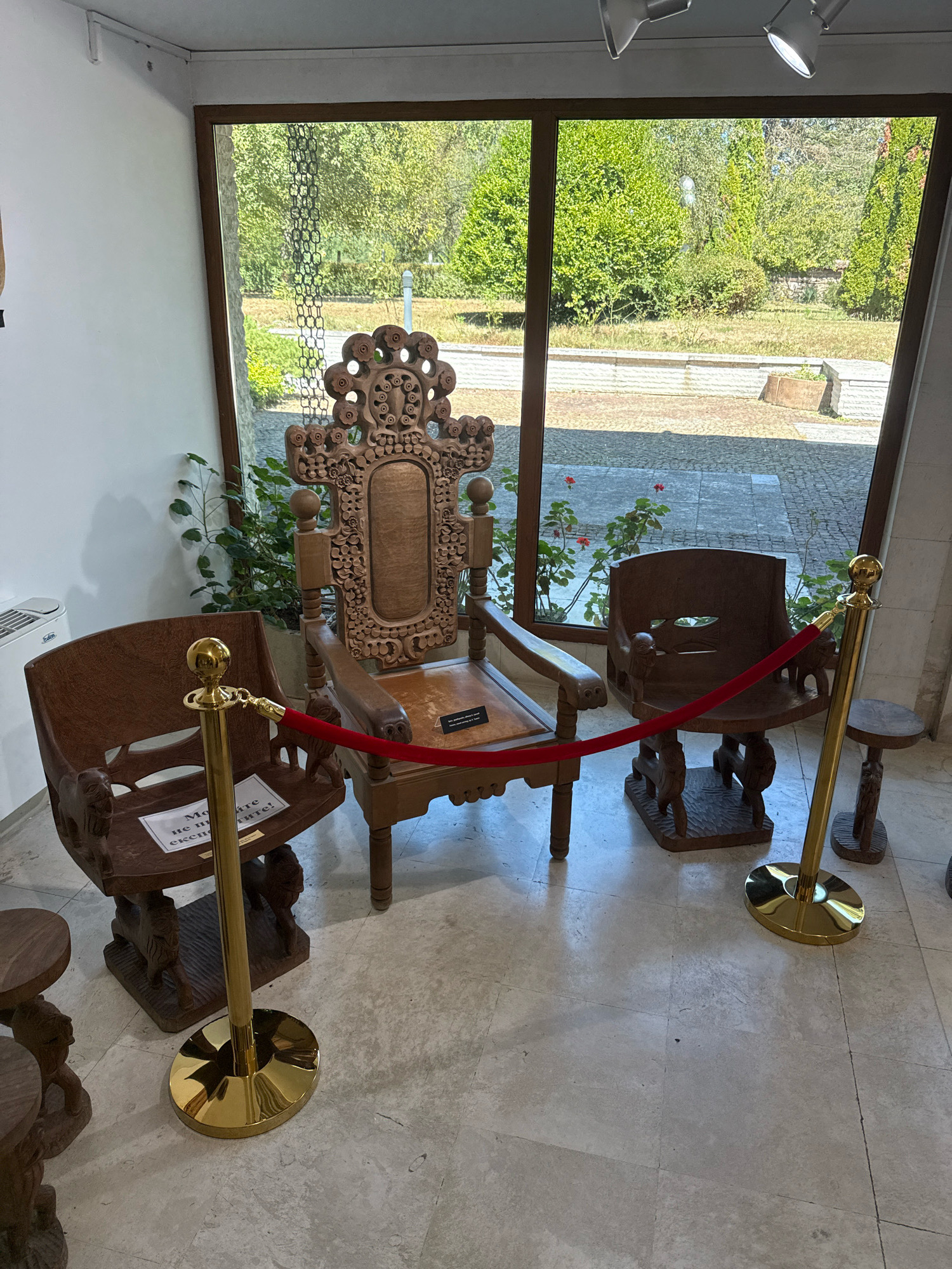 In a brightly lit indoor space, likely a museum or exhibit hall in the city of Pravets, Bulgaria, three intricately carved wooden chairs are arranged. A large, ornate throne-like chair sits centrally, flanked by two smaller, simpler chairs. A red velvet rope, suspended by gold stanchions, separates the viewer from the exhibit. The chairs are positioned on a polished stone floor. Behind them, a large window offers a view of lush green trees and foliage, suggesting an outdoor setting beyond the exhibition space. The overall impression is one of an indoor display of traditional or artisanal woodworking.