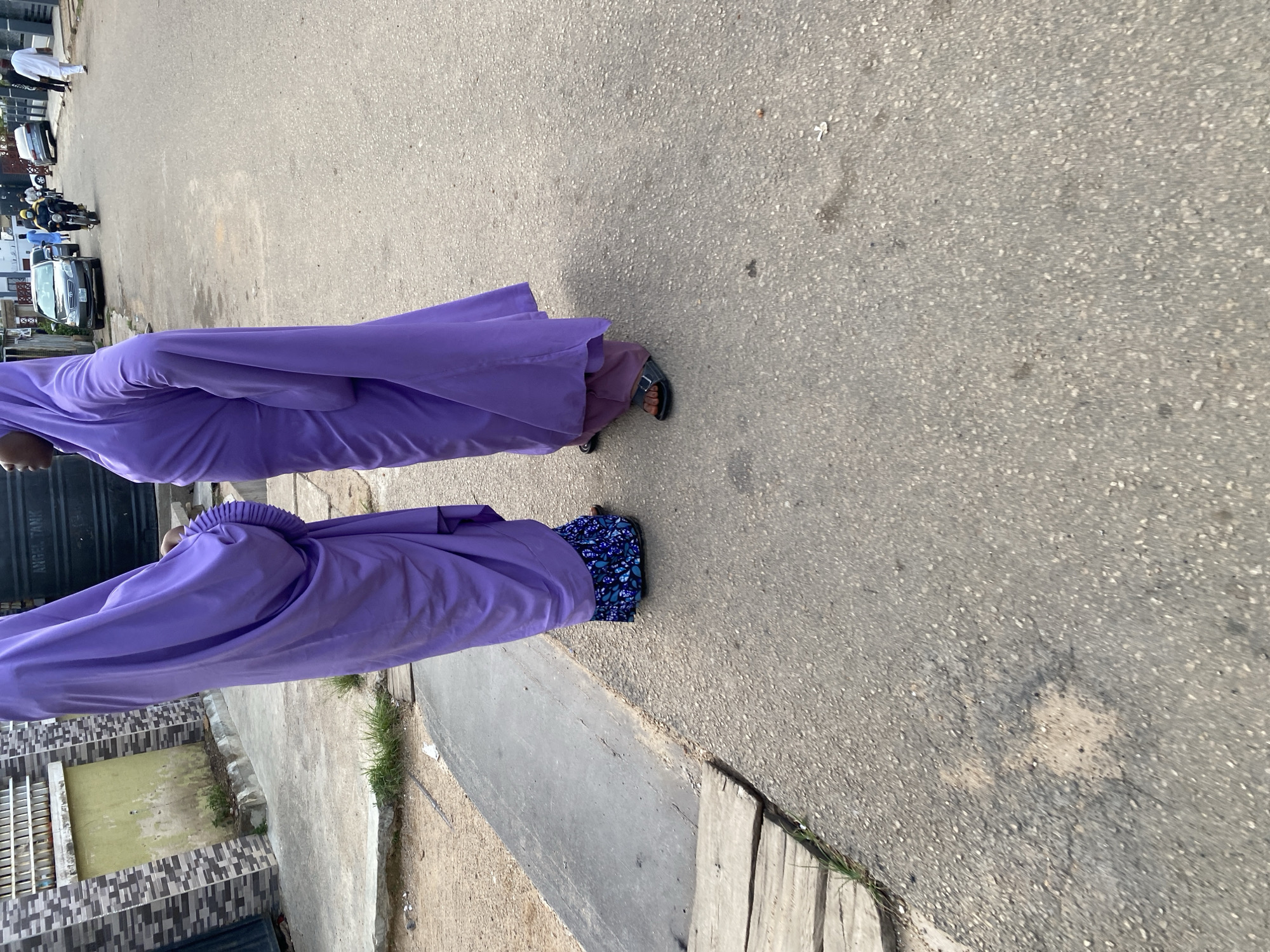 Two women dressed in purple abayas stand on a paved street in Jos, Nigeria. The woman on the left wears a patterned blue skirt visible beneath her abaya, while the woman on the right wears a purple skirt. Both women are wearing sandals. In the background, several cars are parked along the street, and a man on a motorcycle is passing by. A large black water tank is visible on the left side of the image, next to a building with a patterned facade. The street is paved with asphalt and shows signs of wear and tear. The lighting suggests it is daytime, possibly overcast.