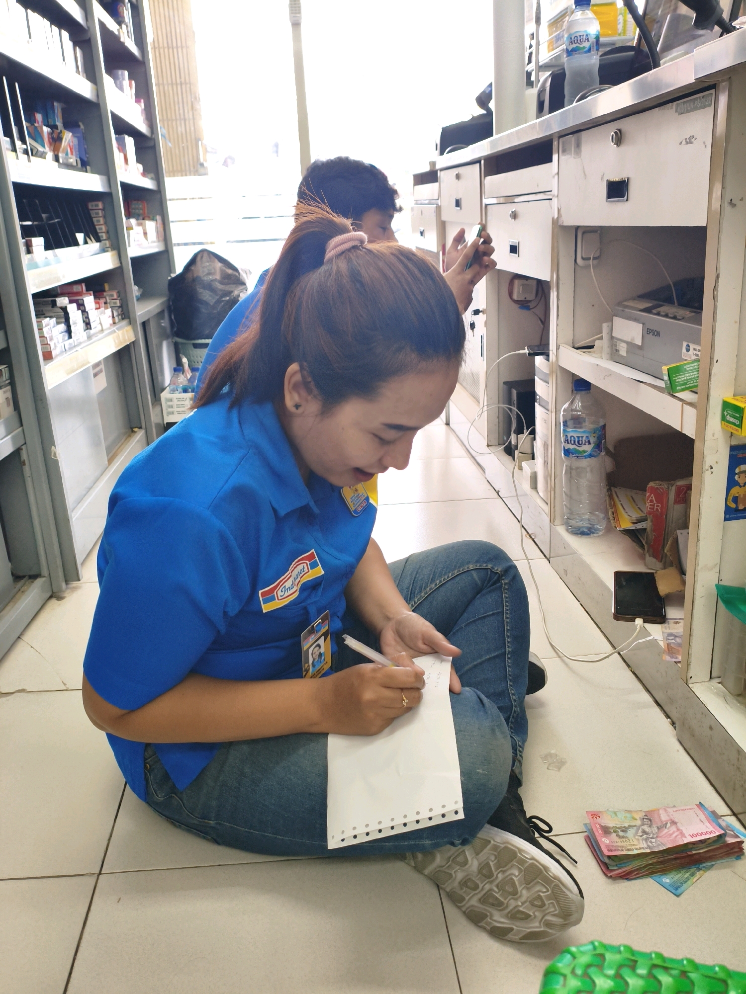 A woman wearing a blue uniform with an "Indomaret" logo is sitting on the tiled floor of what appears to be a convenience store. She is writing on a piece of paper with a pen. She is wearing blue jeans and grey sneakers. To her right, on the floor, is a stack of Indonesian Rupiah banknotes. In the background, there are shelves stocked with products, a counter with drawers, and a bottle of "AQUA" water. Another person is partially visible behind her. The location is identified as the Indomaret in Sumberejo Kulon, Indonesia.